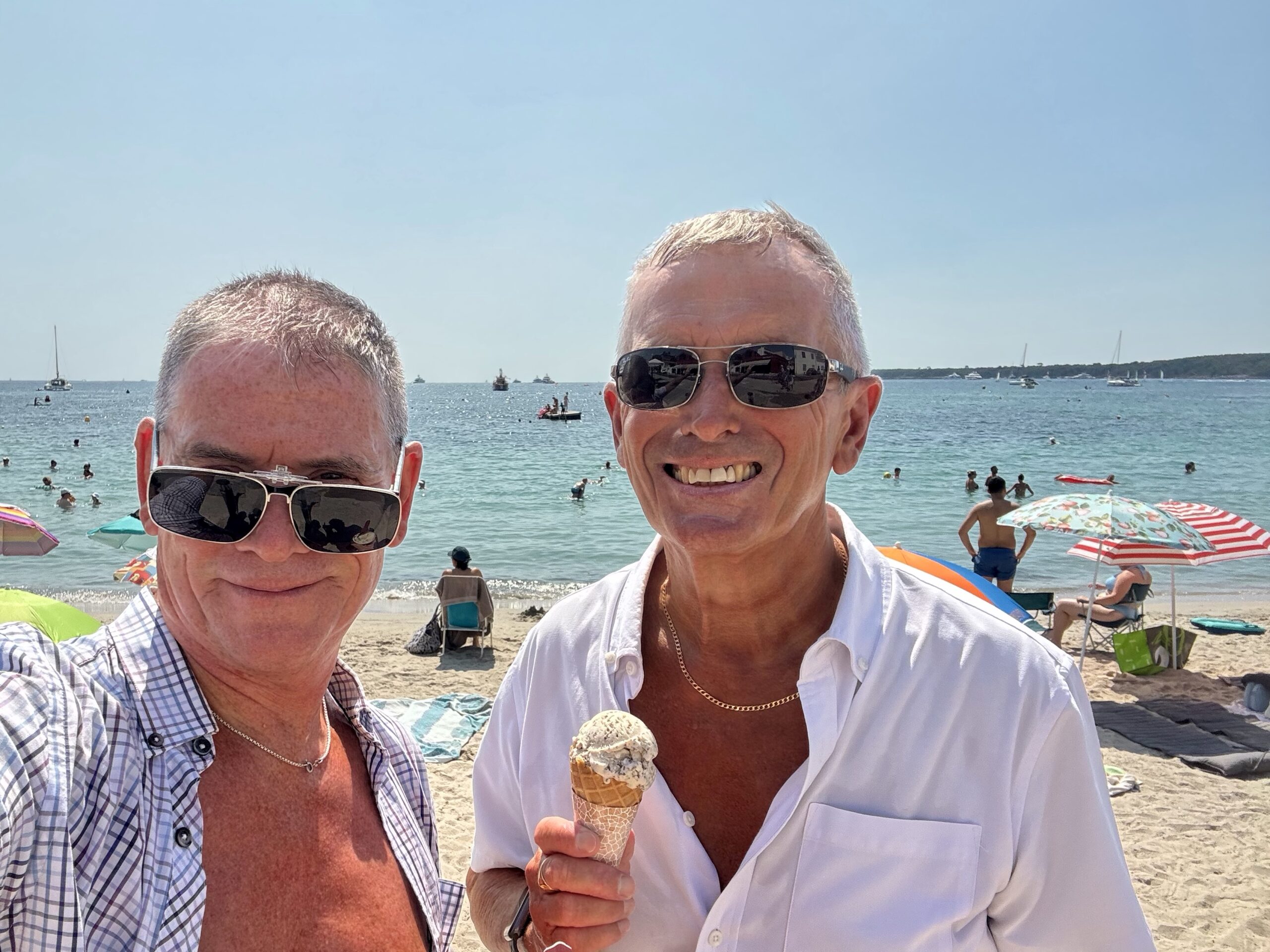 Two men at a sunny beach posing for a photo; one holds an ice cream cone and smiles while the ocean and umbrellas appear behind them.