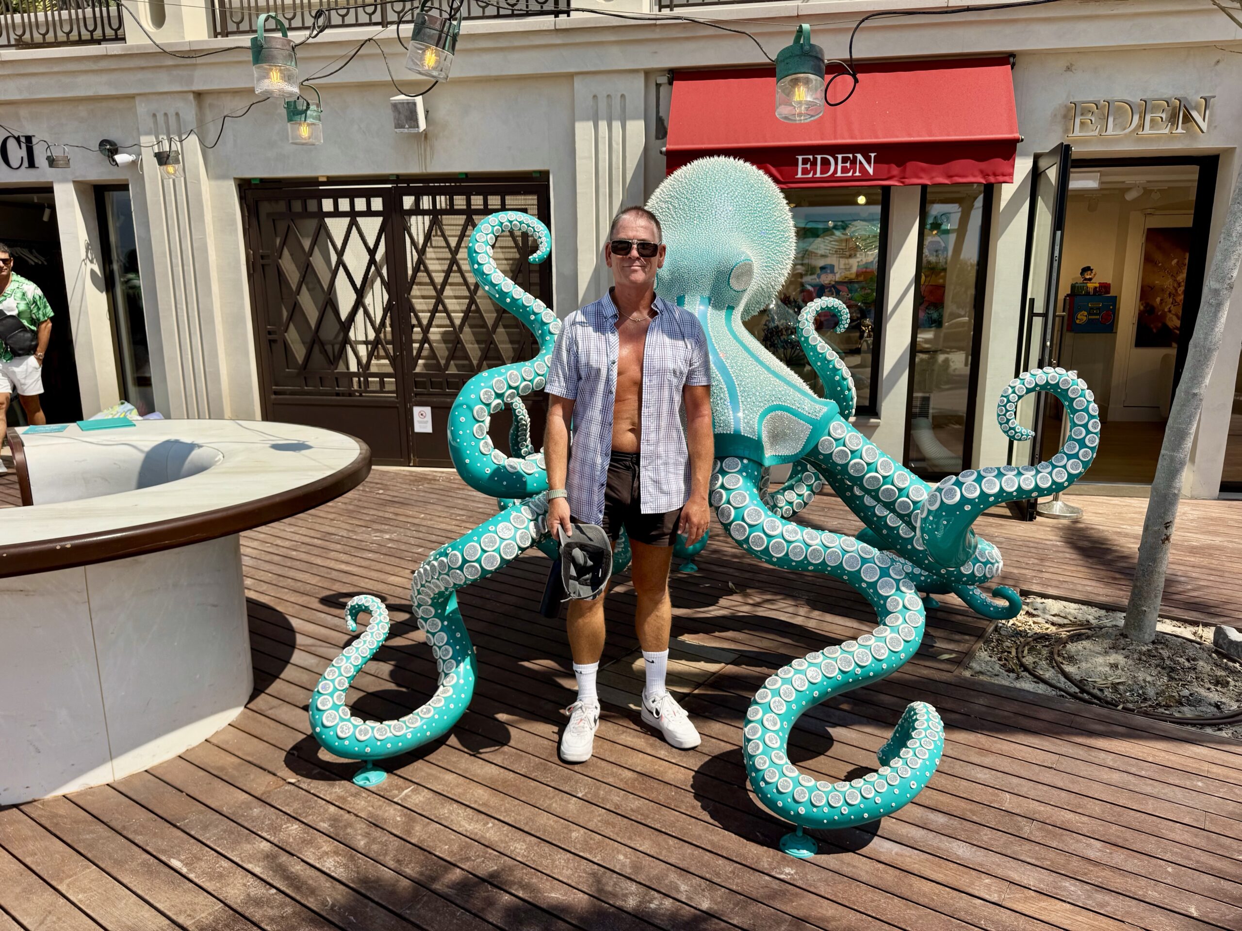 Man posing shirtless under open button-down, standing beside a turquoise octopus sculpture on a wooden deck with an EDEN storefront in the background