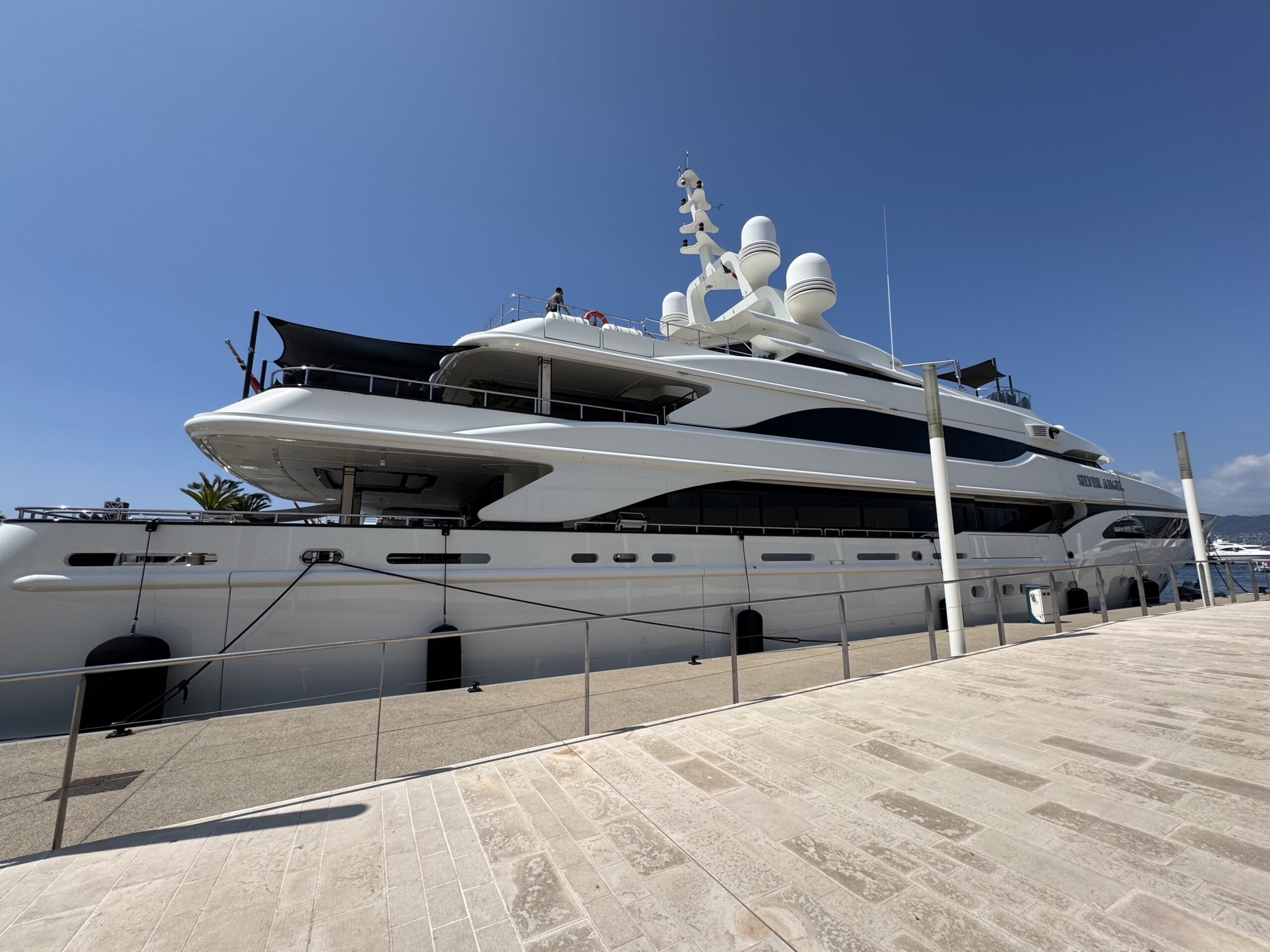 Large white luxury yacht docked at a marina under a clear blue sky, with a person on the upper deck near the railings.