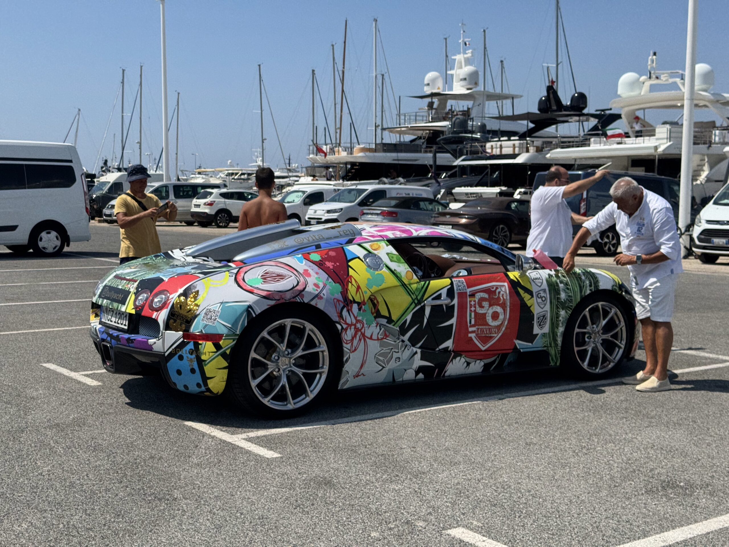 Colorful graffiti-covered sports car in a marina parking lot with people and luxury yachts in the background.