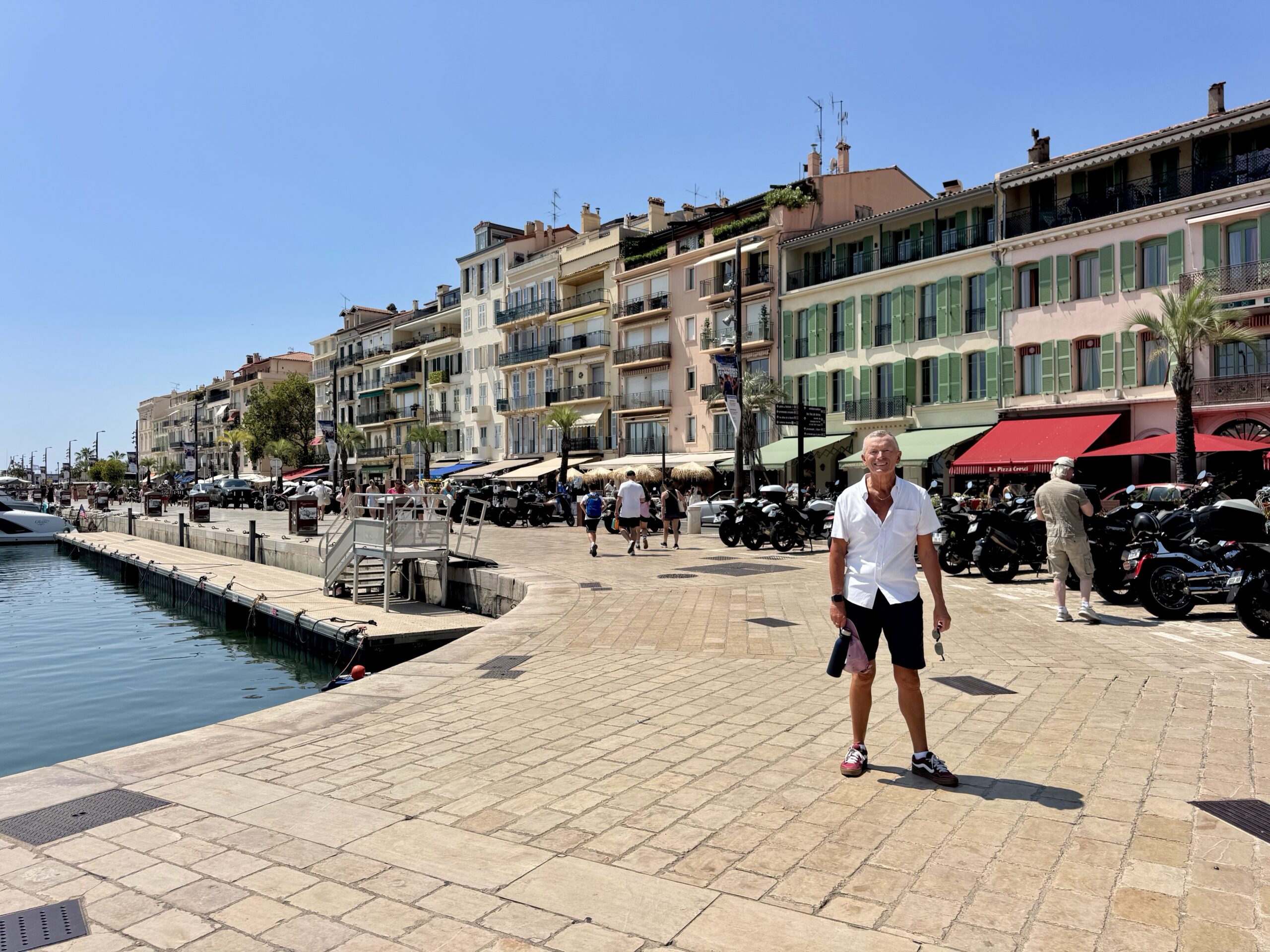 Sunlit seaside promenade with pastel buildings, outdoor cafes, and people strolling along the harbor.