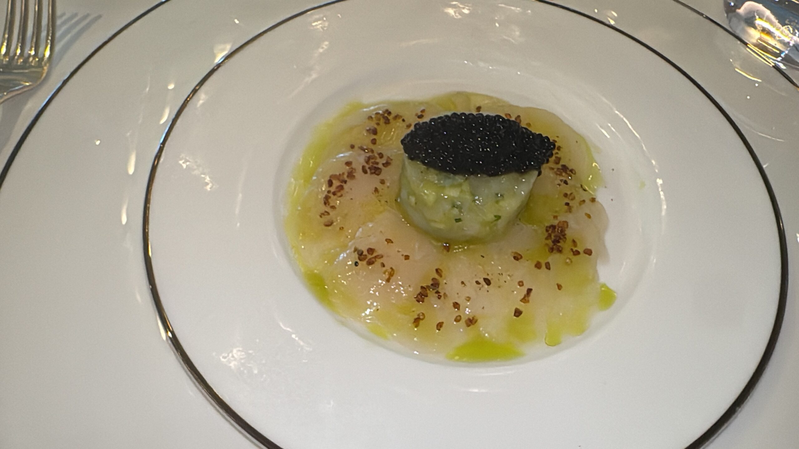 Single seared scallop topped with black caviar on a green purée, finished with olive oil and scattered seeds on a white plate with a decorative rim.