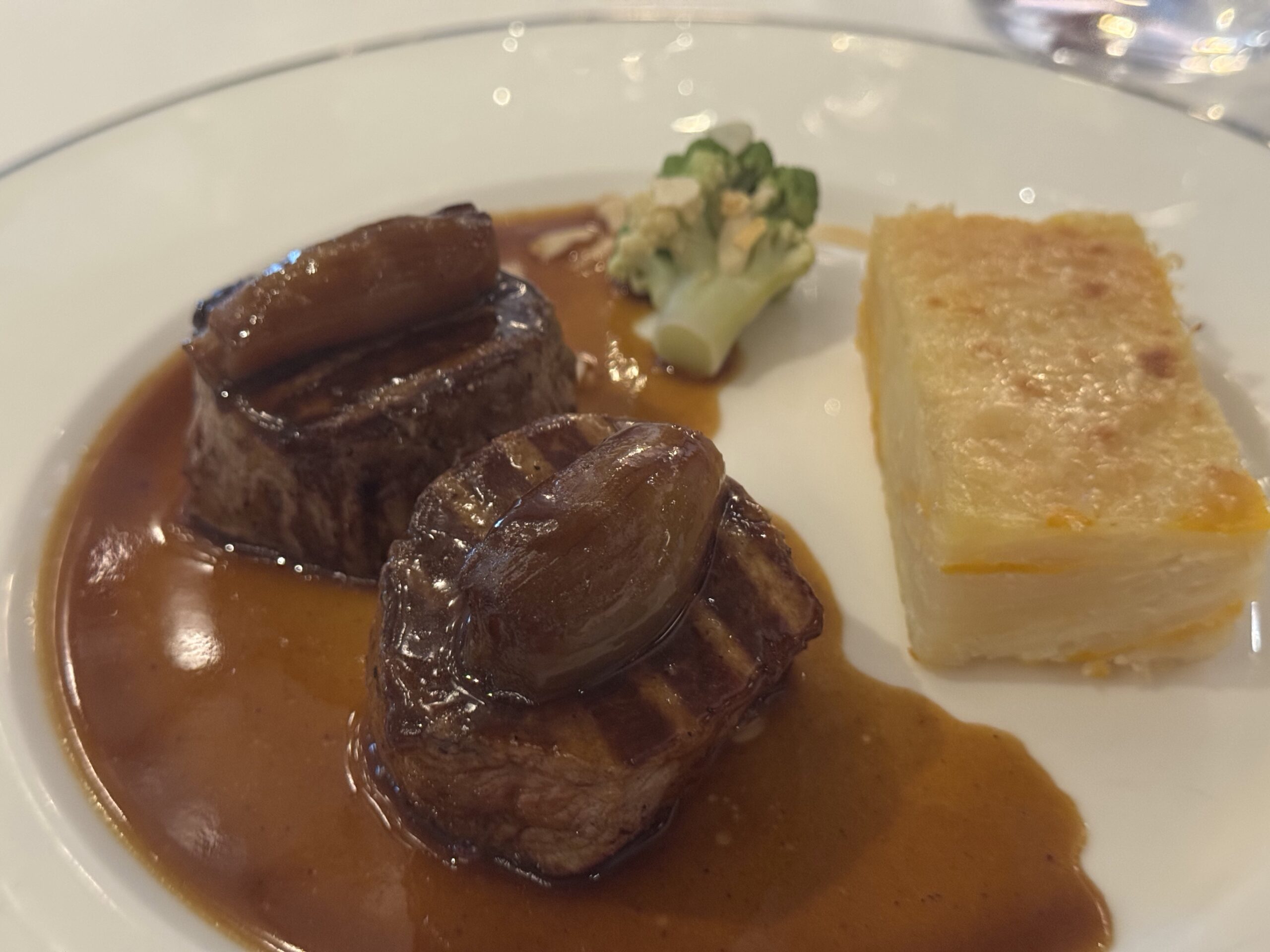 Plate featuring braised beef in brown gravy with a broccoli floret and almond topping, and a rectangular potato gratin on the side.