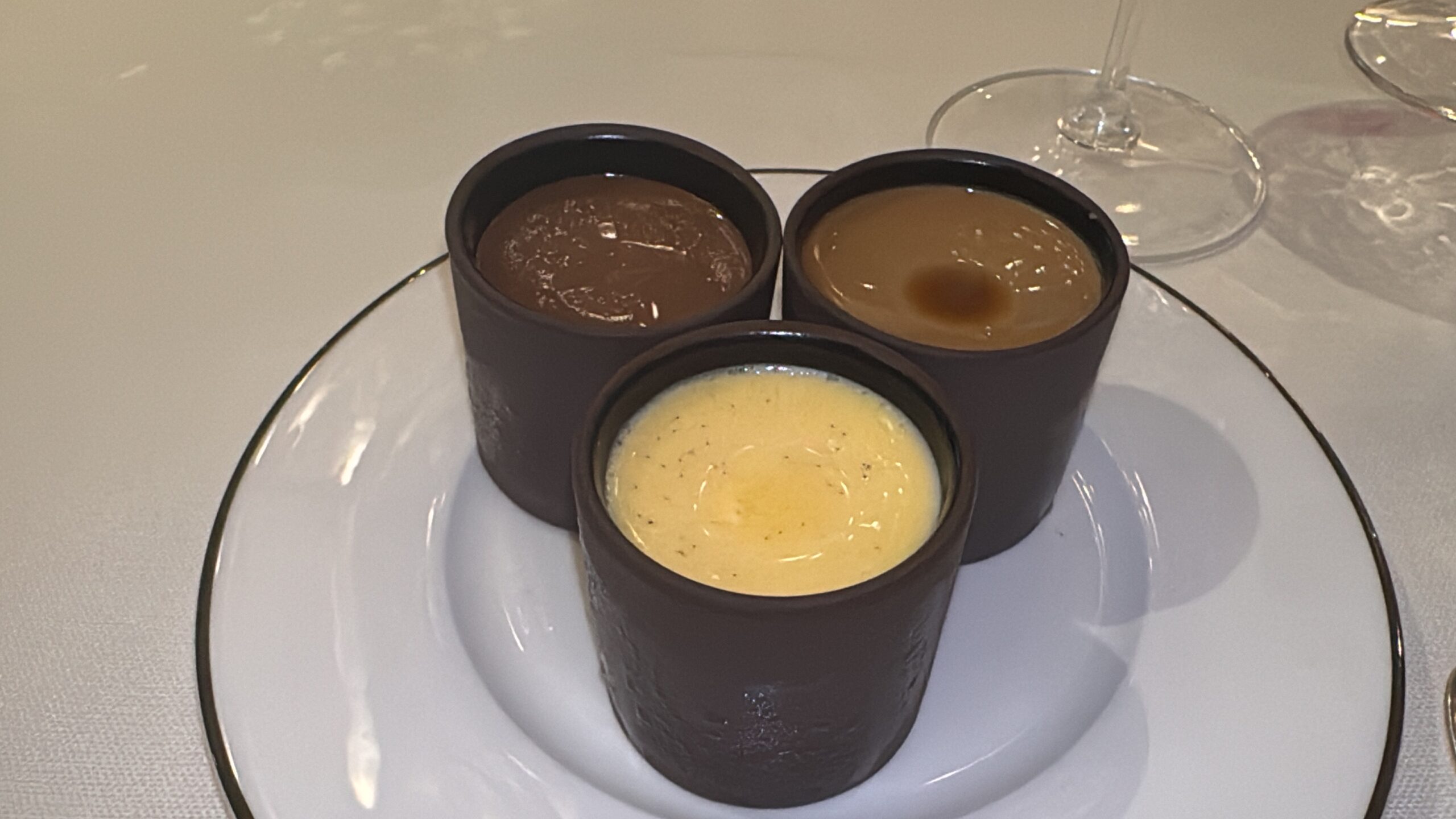 Three small dark bowls with chocolate, caramel, and vanilla sauces arranged on a white plate with a wine glass nearby.