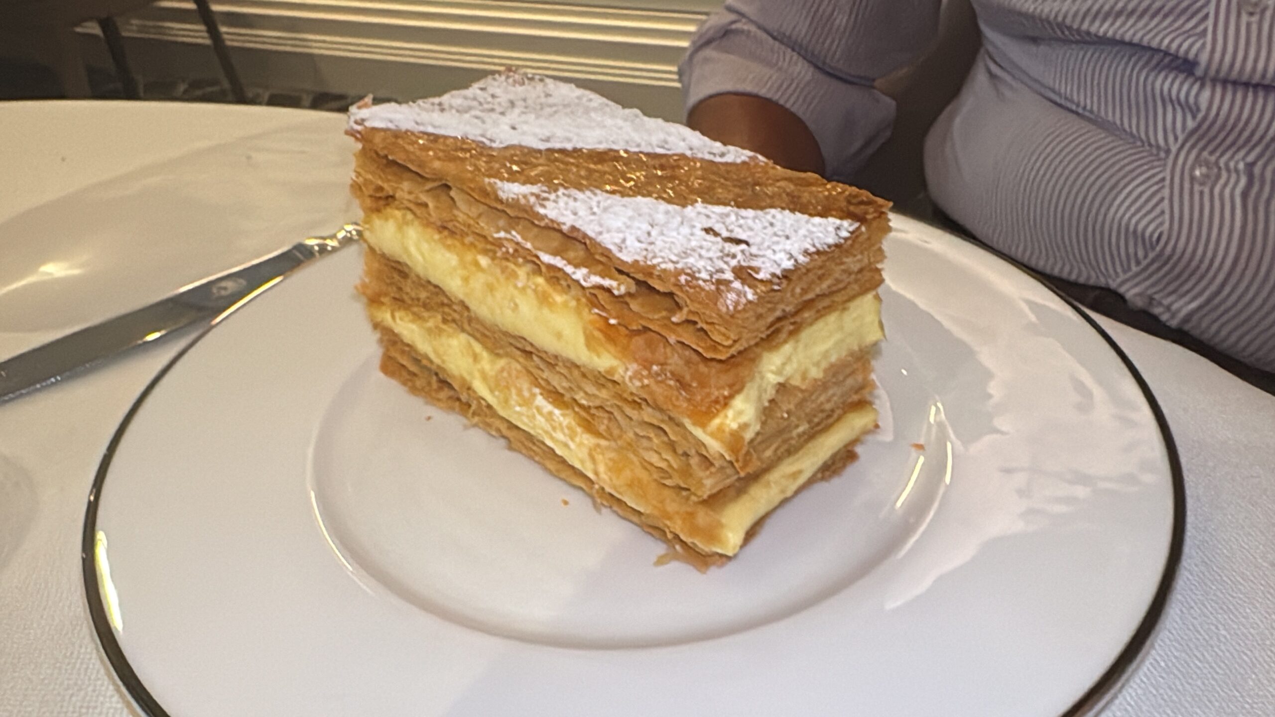 Slice of mille-feuille pastry with powdered sugar on a white plate, a knife nearby.