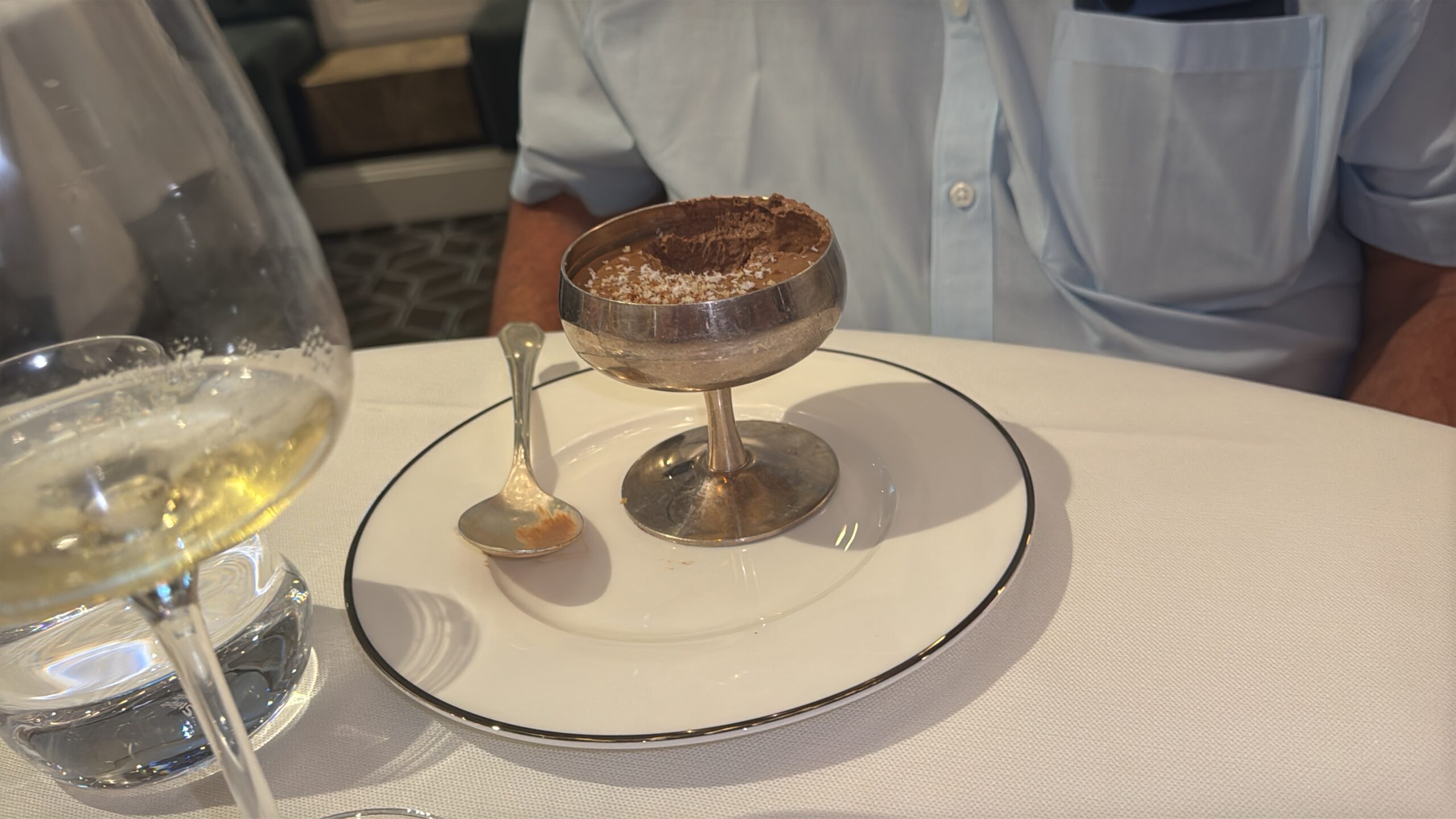 Chocolate mousse dessert topped with coconut flakes served in a silver coup glass on a white plate at a formal table setting.
