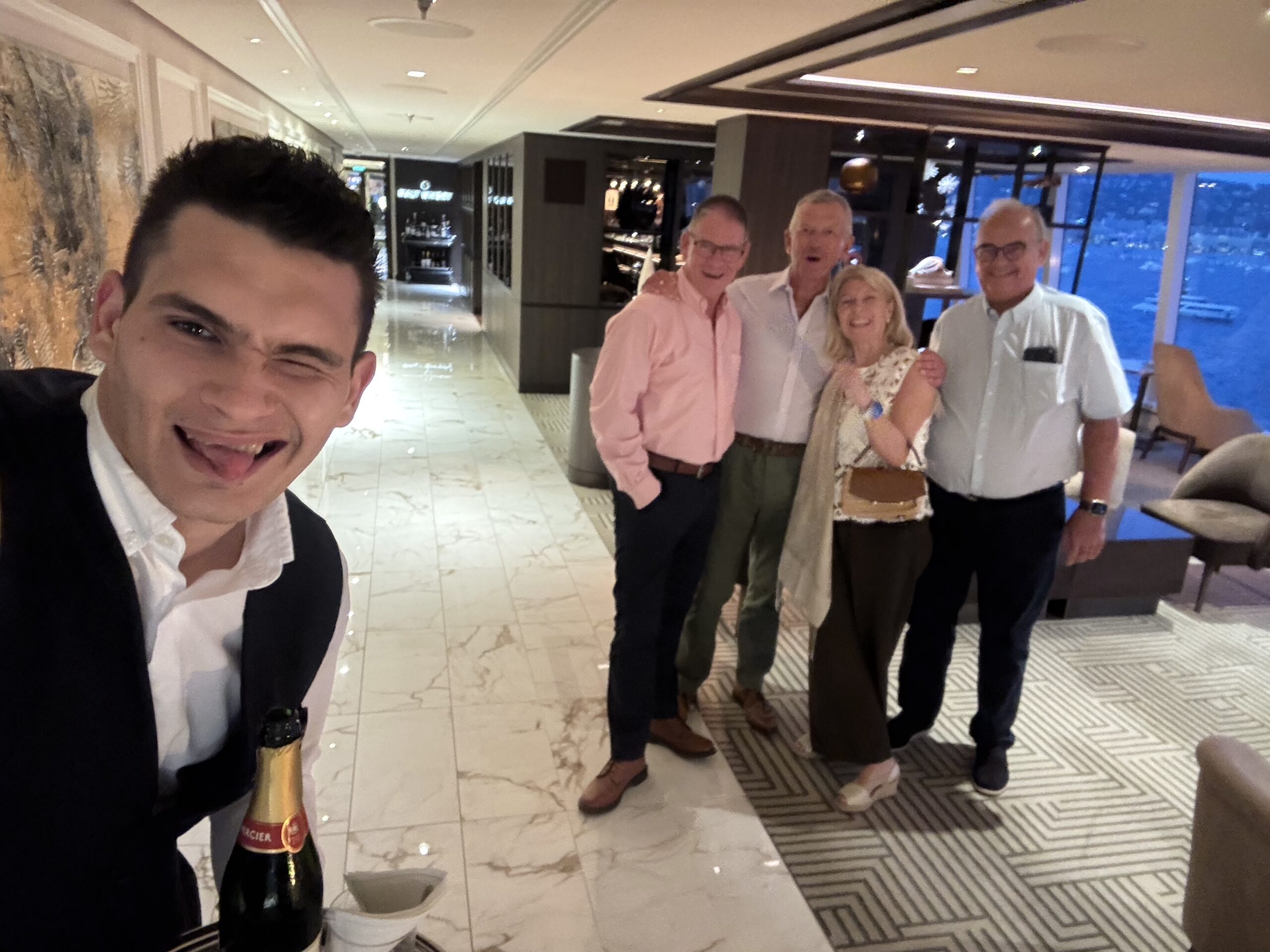 Man in a suit taking a selfie with a champagne bottle, four friends smiling in a stylish hotel lobby behind him.