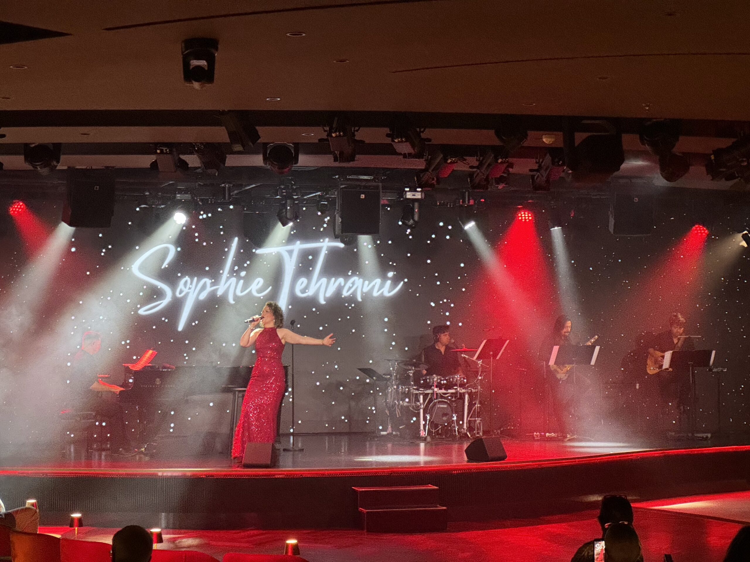 Singer in a sparkling red gown performs on a lit stage with a band; backdrop reads 'Sophie Tehrani'.