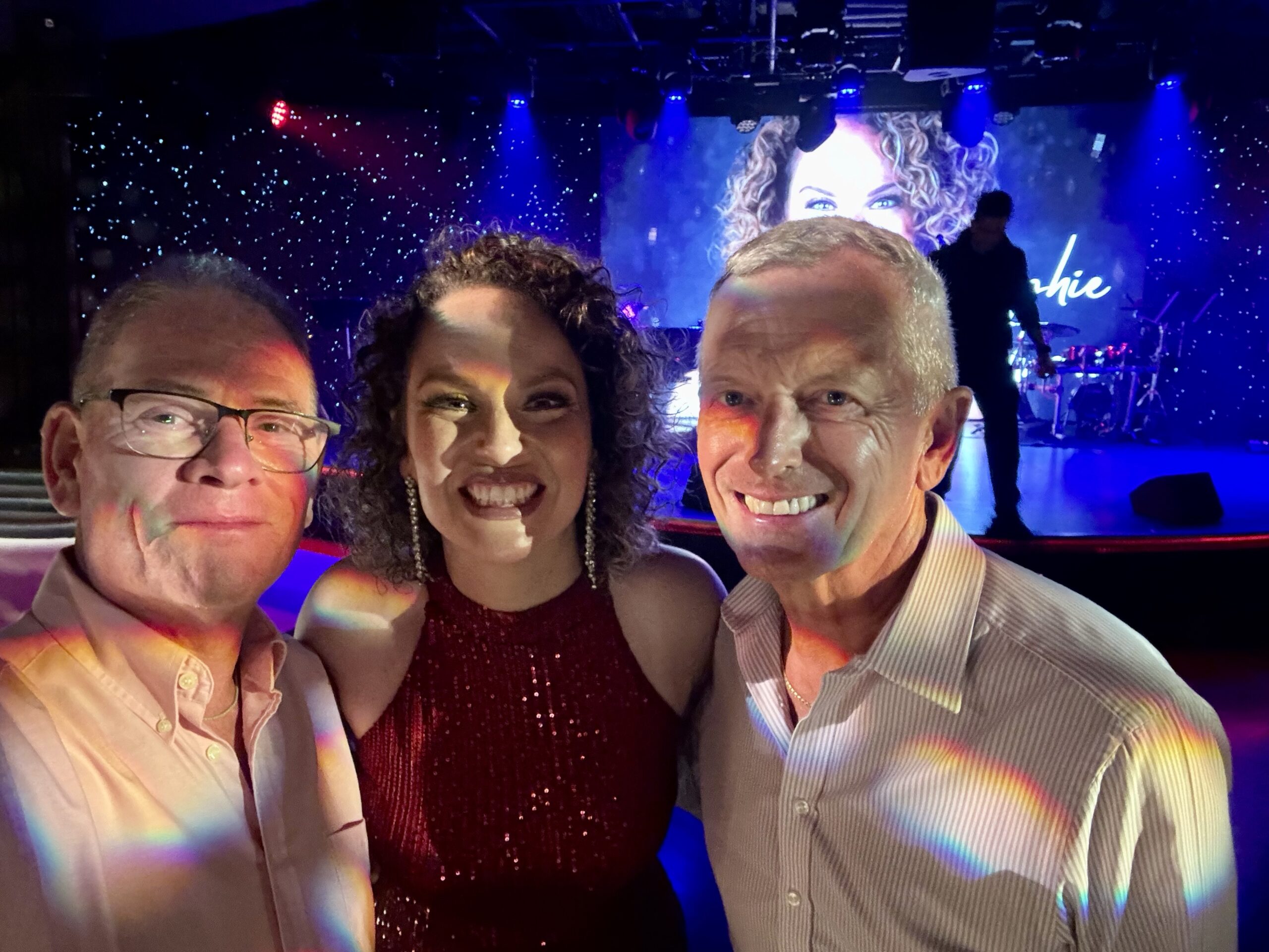 Three friends smile for a selfie on a music venue floor with a stage and blue lights in the background. Colorful rainbow light streaks appear on their clothes.