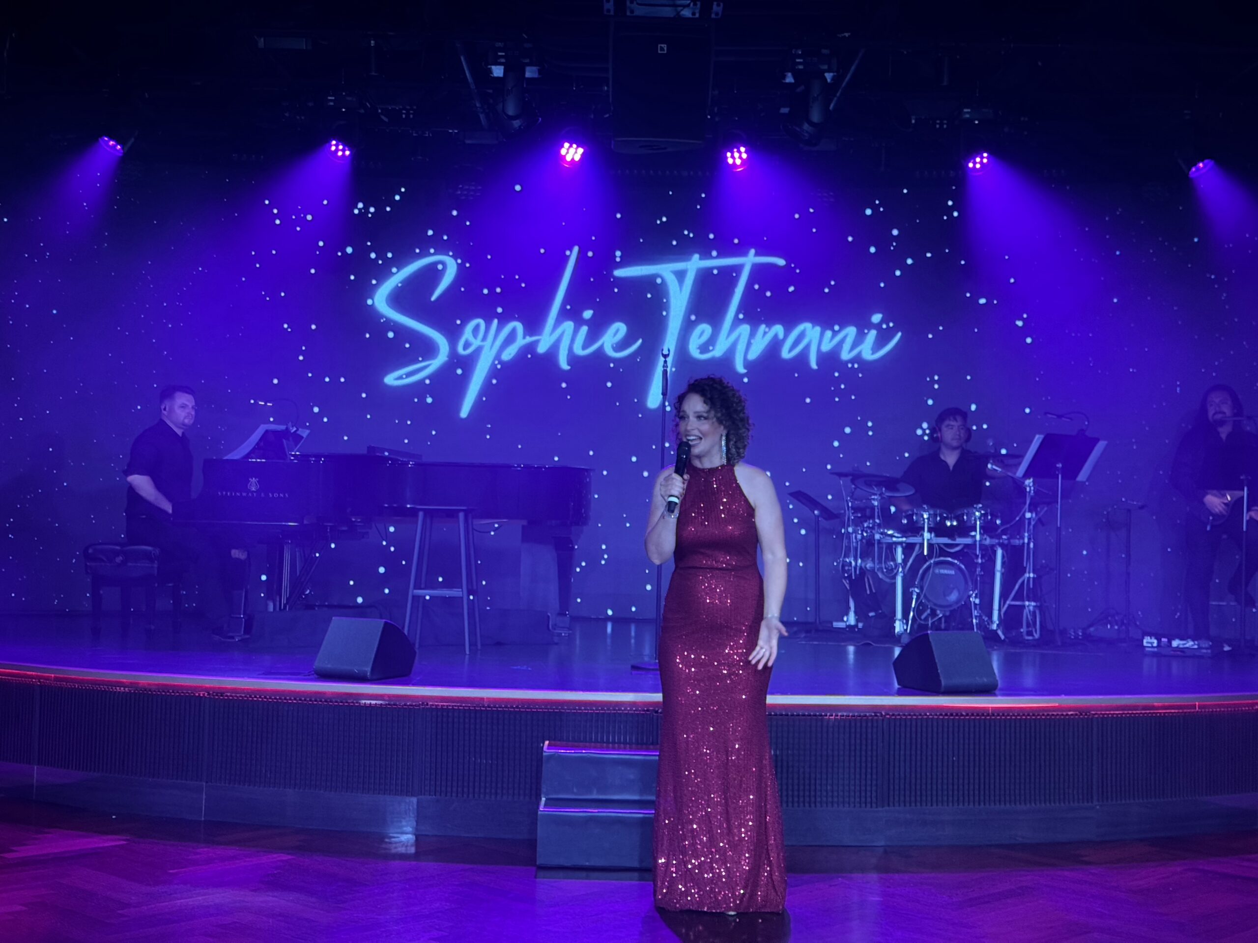 Singer center stage in a red sequined gown performing under purple lights, with a backdrop showing the name Sophie Tehrani and a live band.