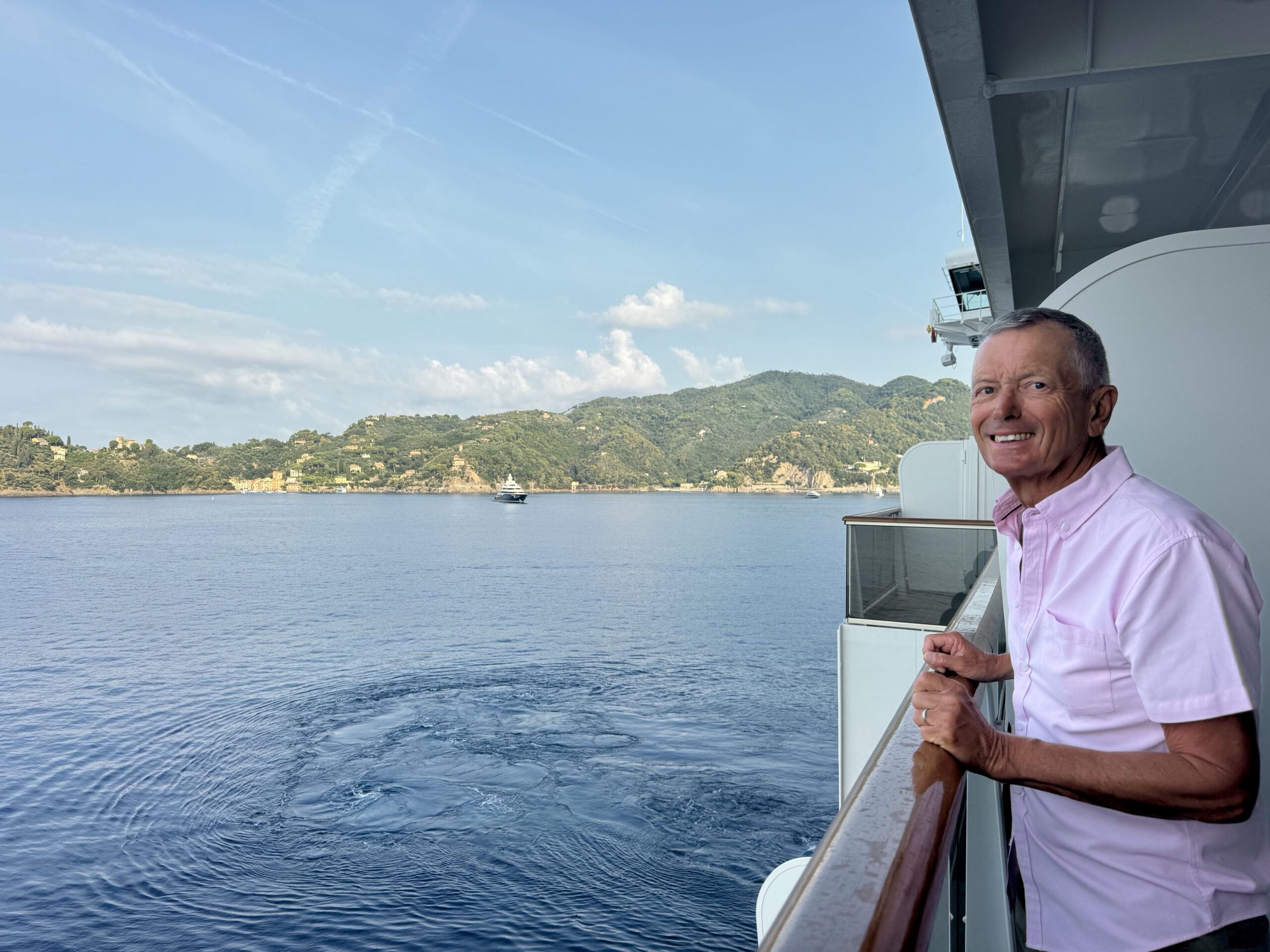 Smiling man in a pink shirt leaning on a ship balcony railing with blue sea and distant hills behind him.