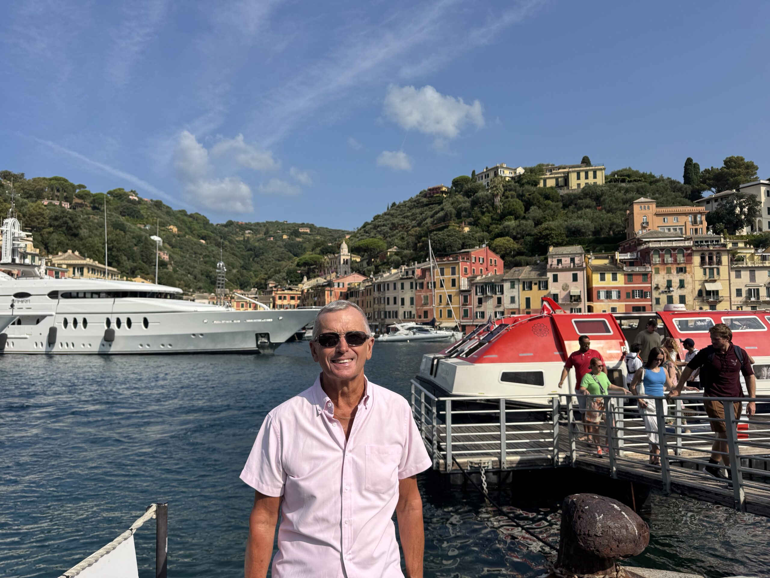 Smiling man in a pink shirt stands on a marina dock with colorful hillside buildings in the background.