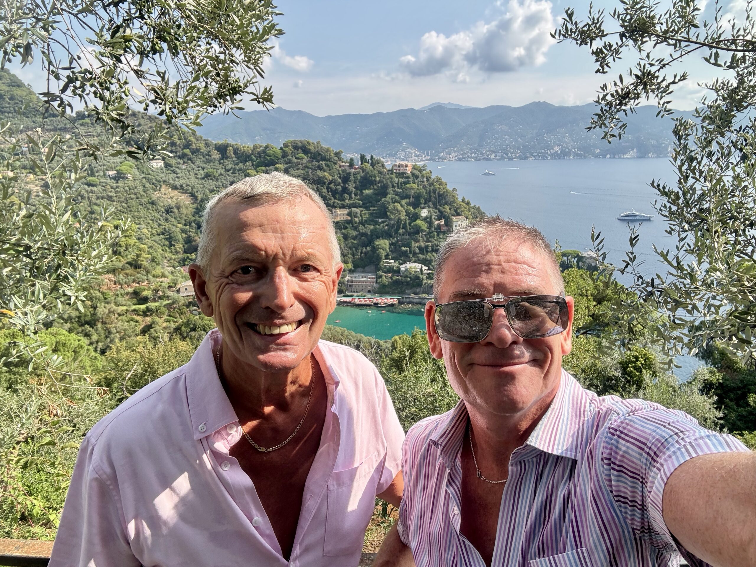 Two men smiling for a selfie with olive trees framing a hillside town and blue sea in the background.