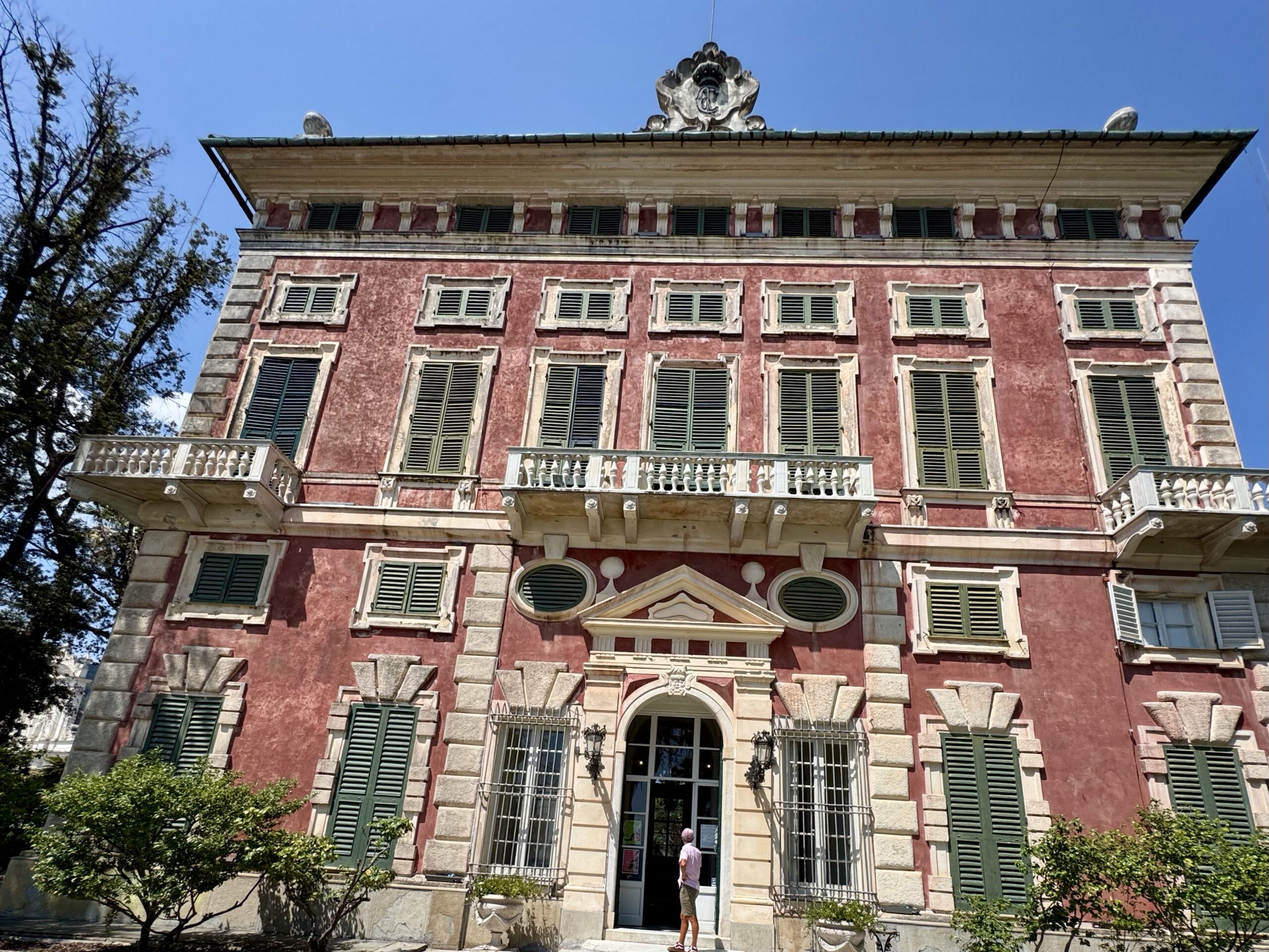 Historic red stucco building with green shuttered windows, decorative stone frames, and balconies against a clear blue sky.