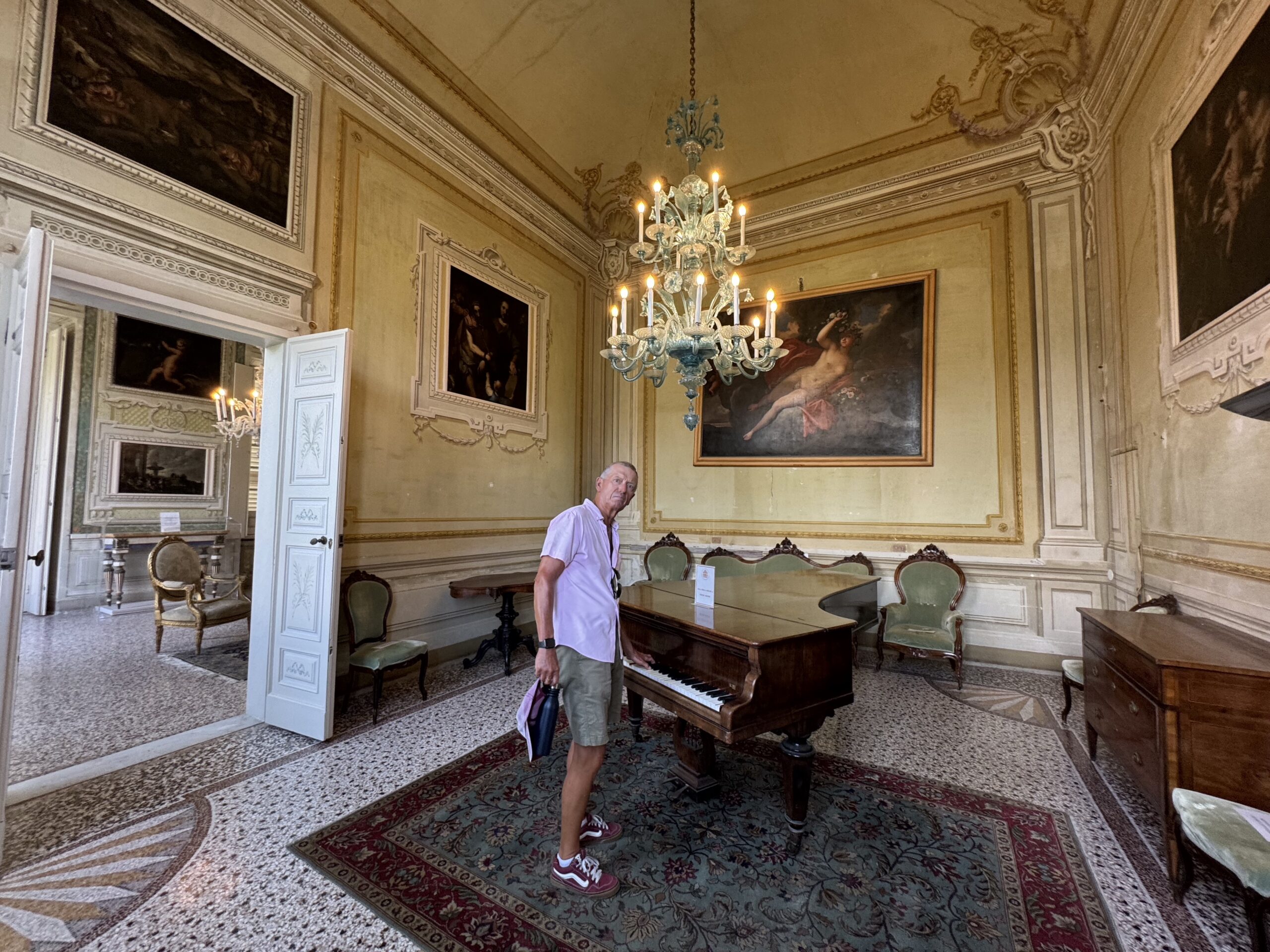 Man standing beside a grand piano in an ornate, gold-trimmed room with a chandelier and large framed paintings.