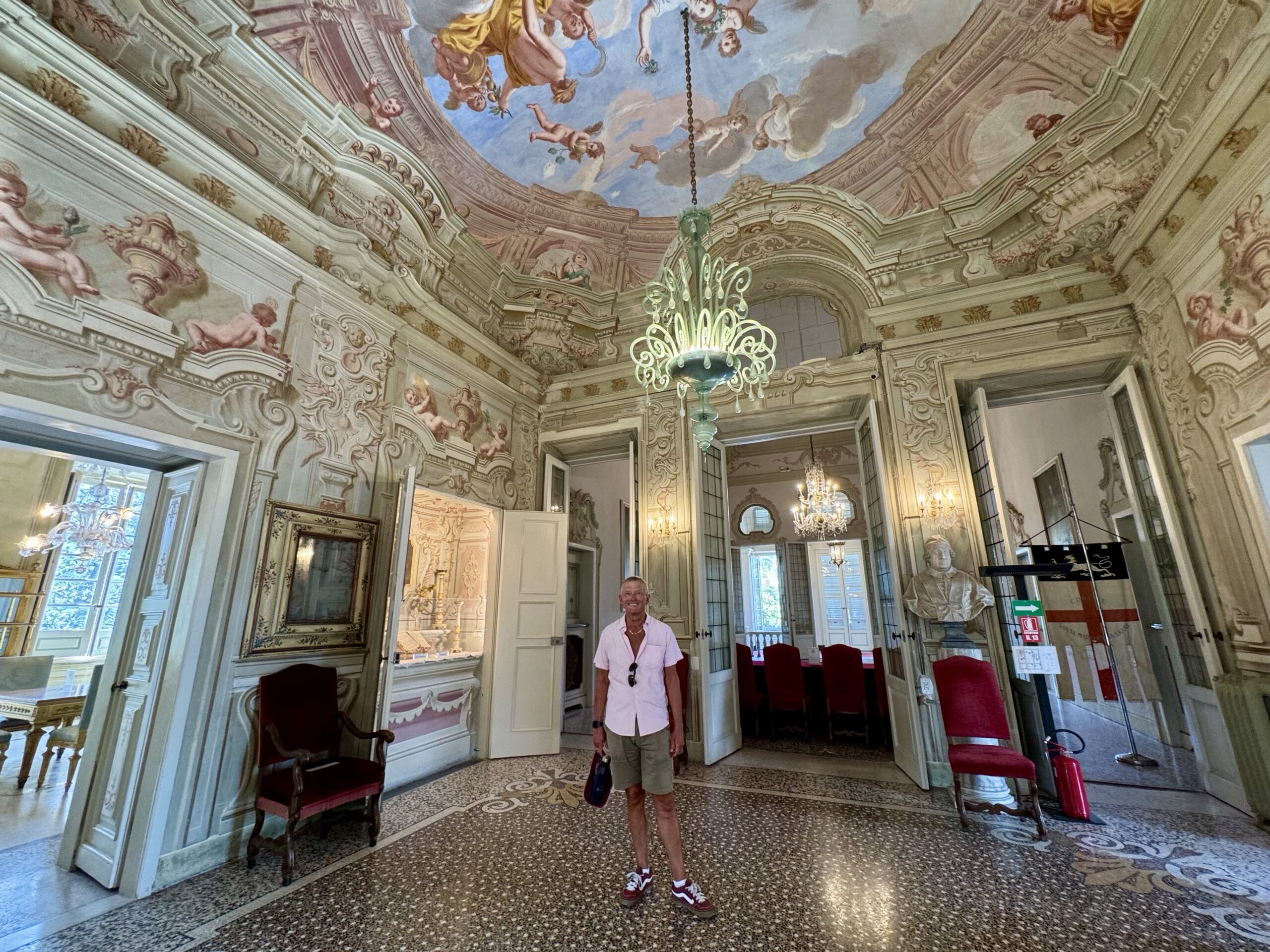 Luxurious rococo interior with a painted ceiling and ornate walls; a man in a light shirt stands in the hall.