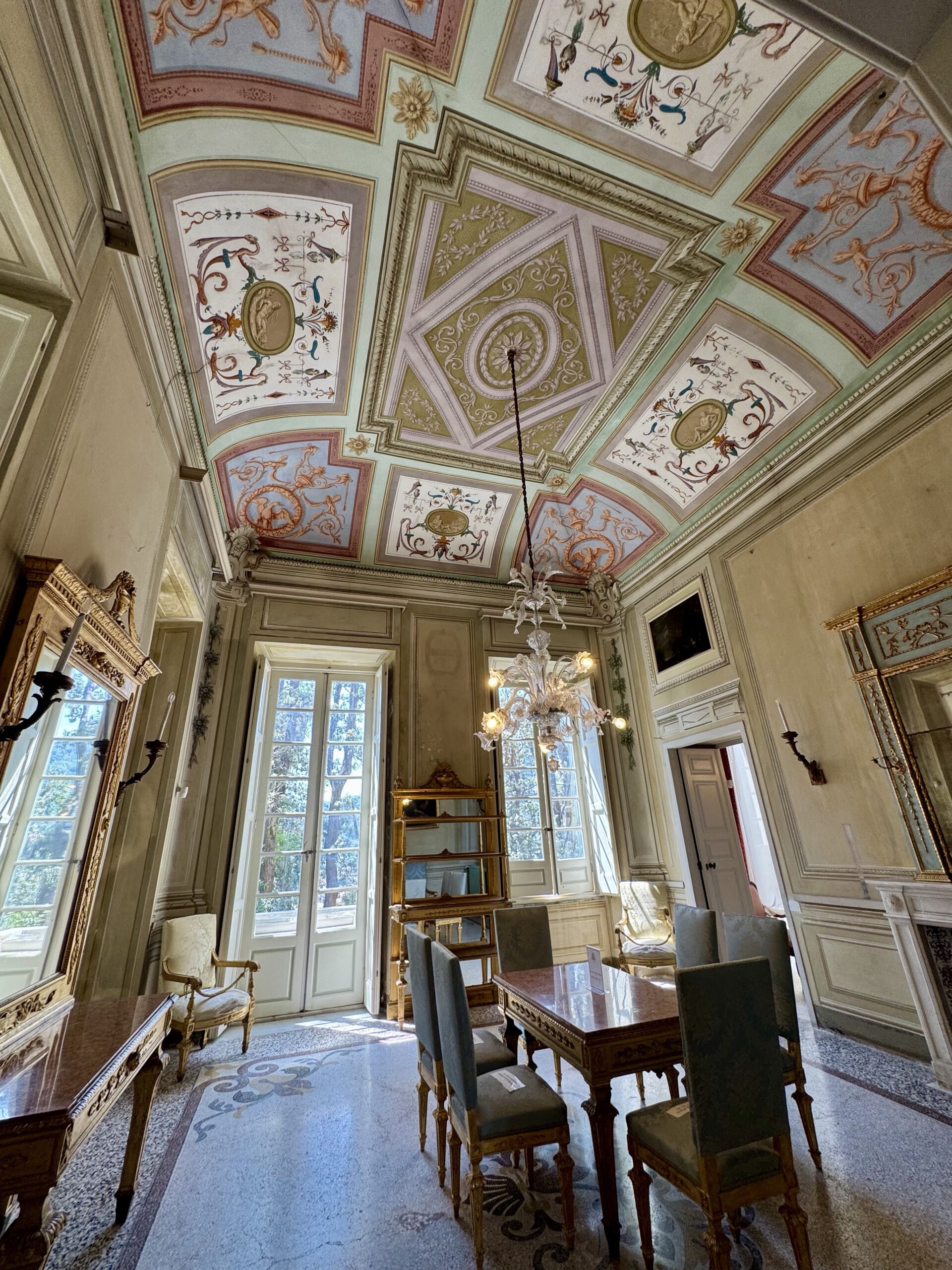 Ornate ceiling with colorful fresco panels above a dining room table and chairs, chandelier hanging center.
