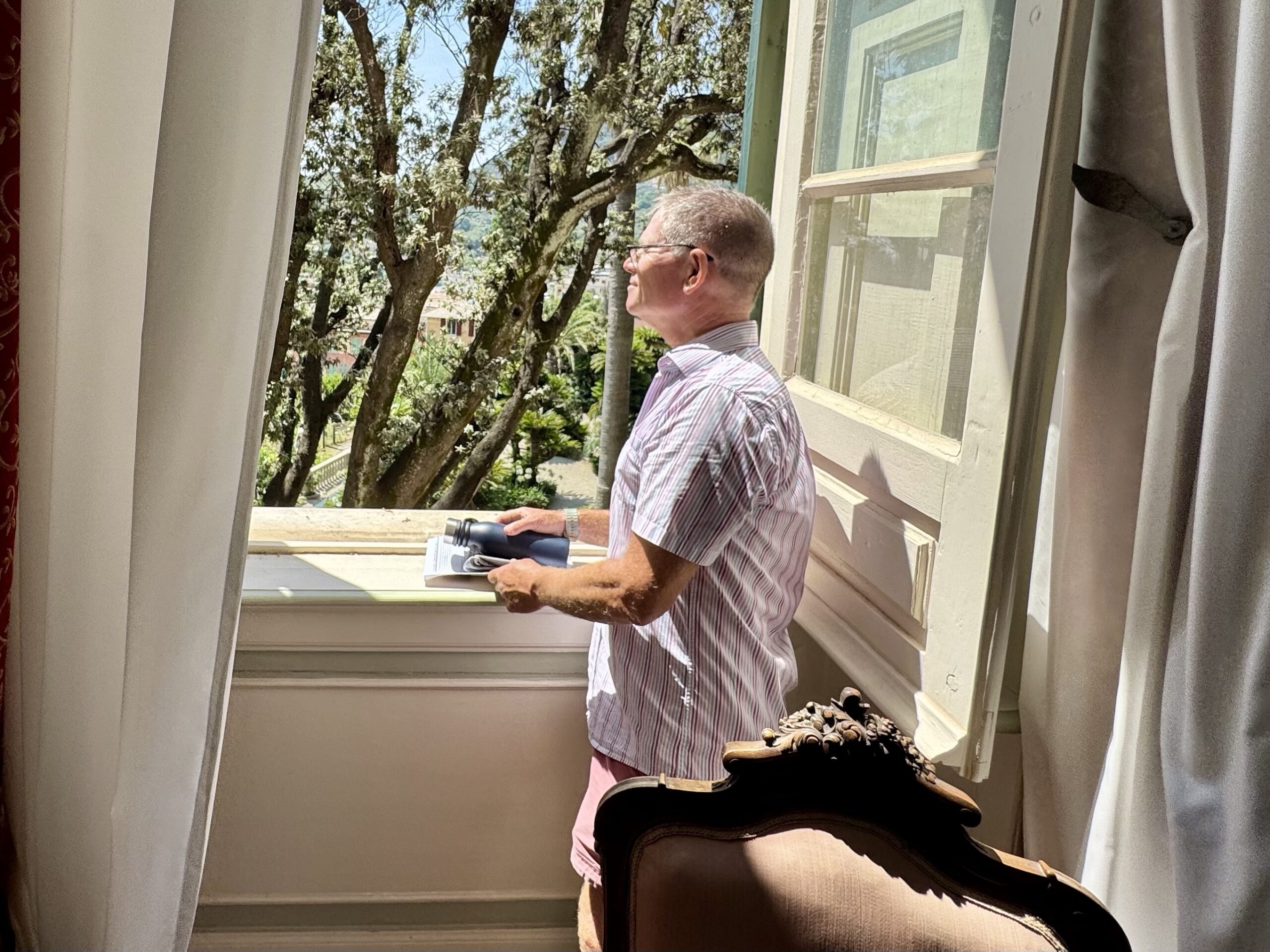 Man in a striped shirt and glasses stands at an open window, holding binoculars and a booklet, looking outside.