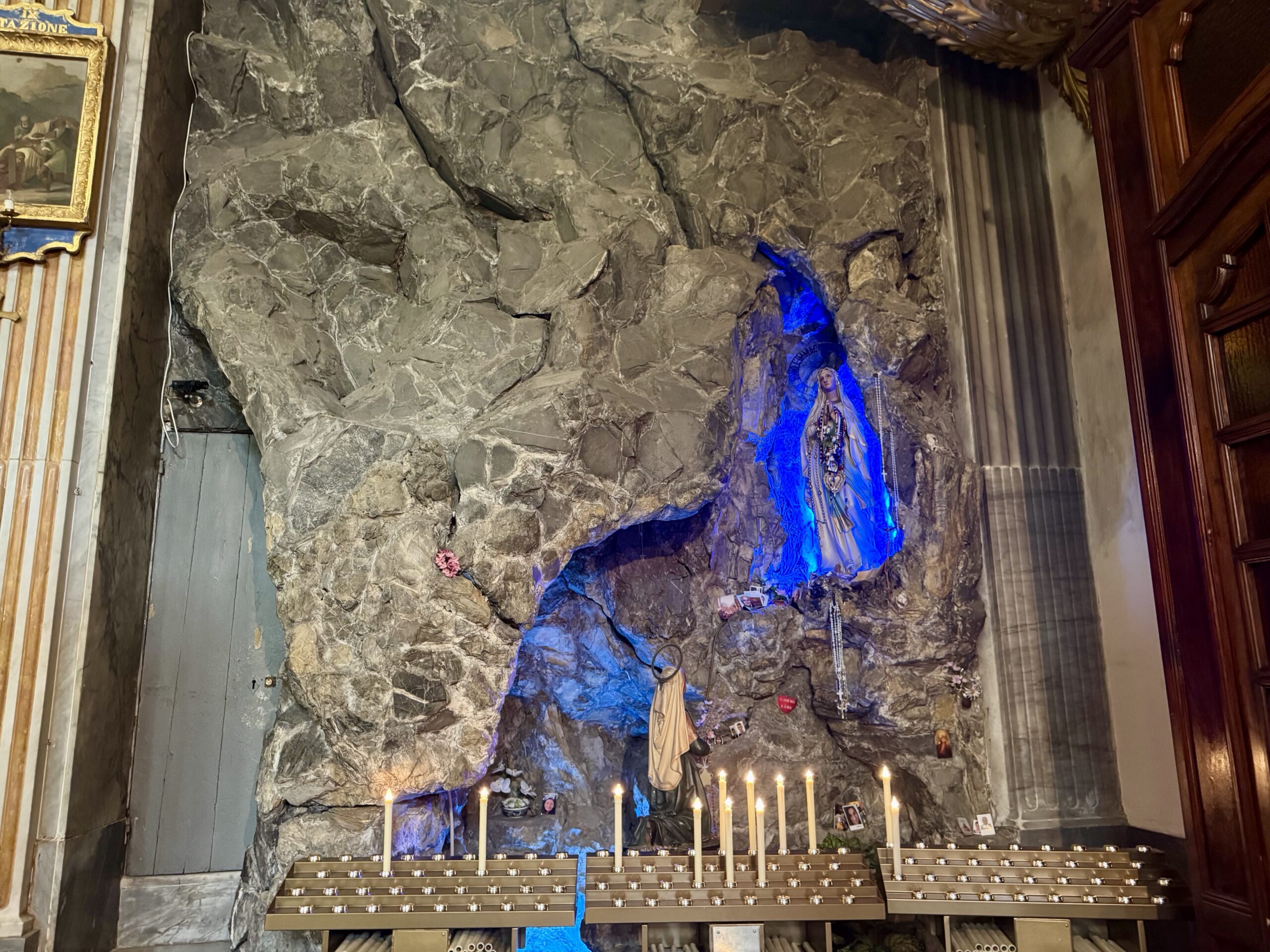 Stone grotto shrine with a statue of Mary (Our Lady) on a rocky wall, blue lighting, candles, and pews in front of the altar area.