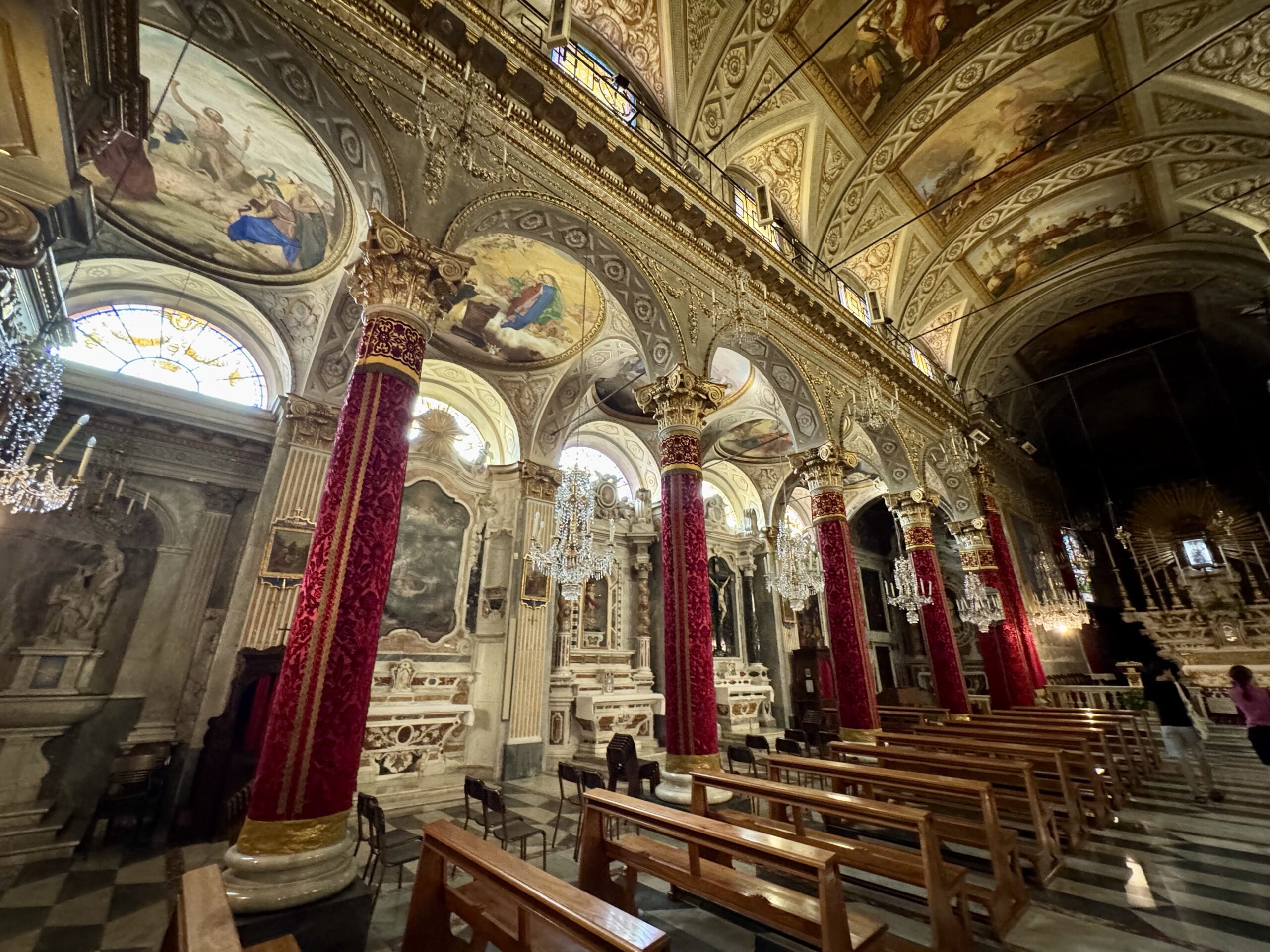 Ornate church interior with gilded arches, frescoed ceilings, chandeliers, and red velvet-covered columns lining wooden pews.