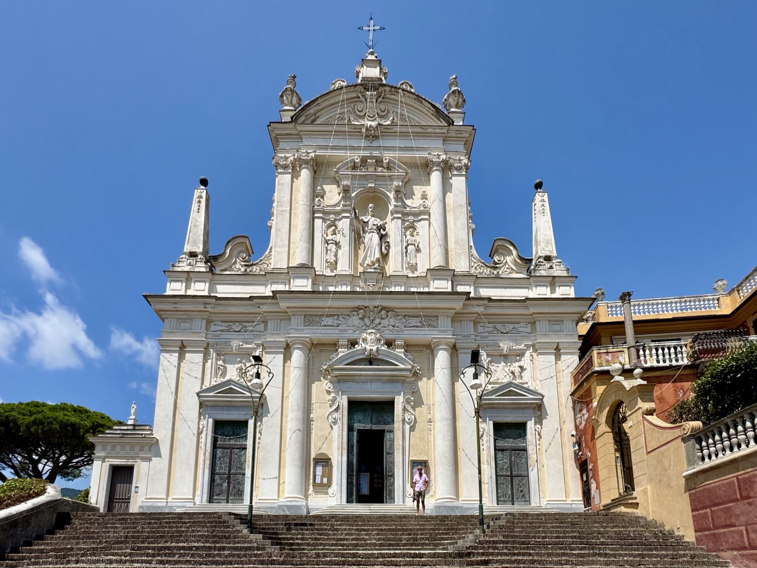 Ornate white church facade with statues, columns, and a decorative pediment, reached by wide stone steps against a clear blue sky.