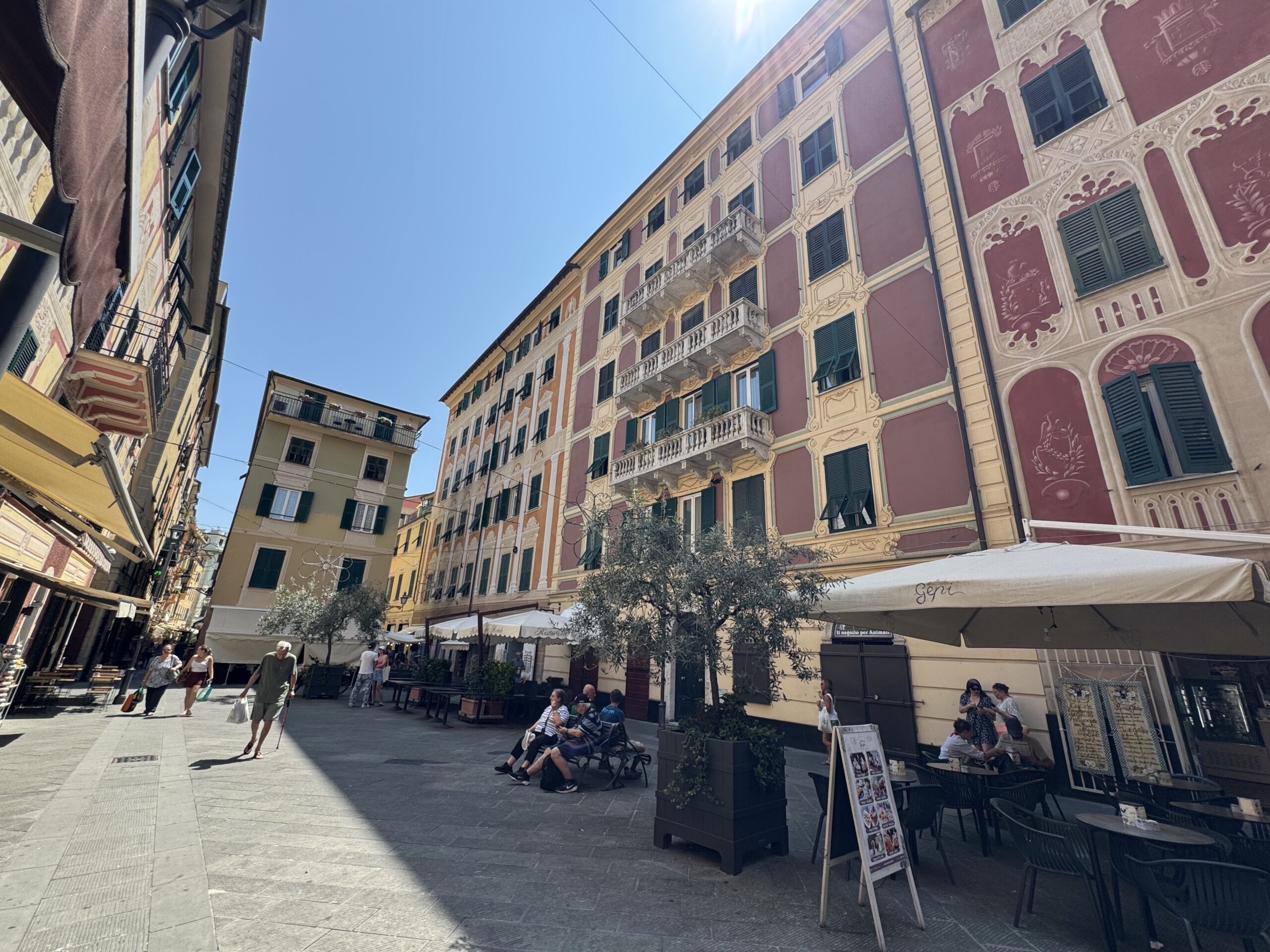 Pedestrian street in a European town with ornate pastel buildings, balconies and green shutters, and outdoor cafe seating under umbrellas.