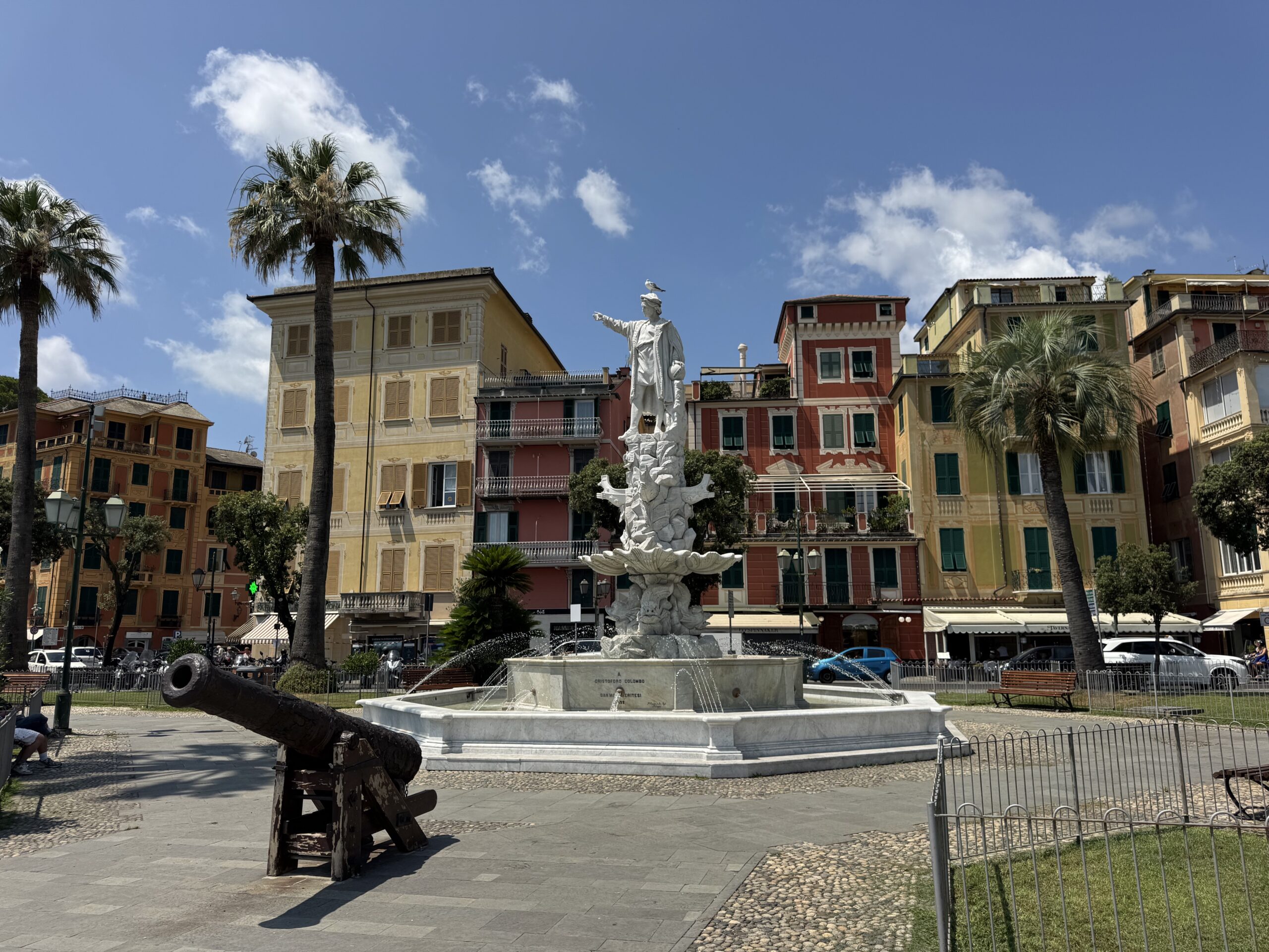 Bright urban square with a white marble statue fountain, palm trees, and pastel buildings under a blue sky.