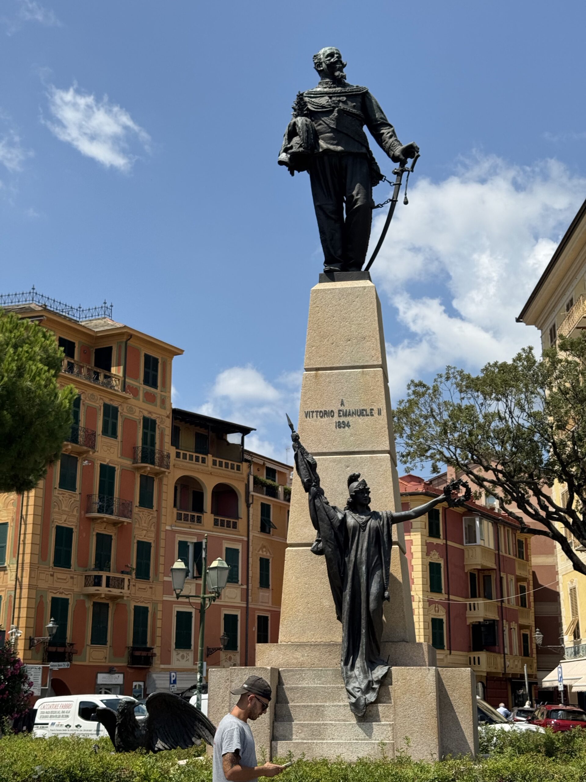 Bronze statue of Vittorio Emanuele II on a tall pedestal in a square, holding a sword, with colorful buildings in the background.