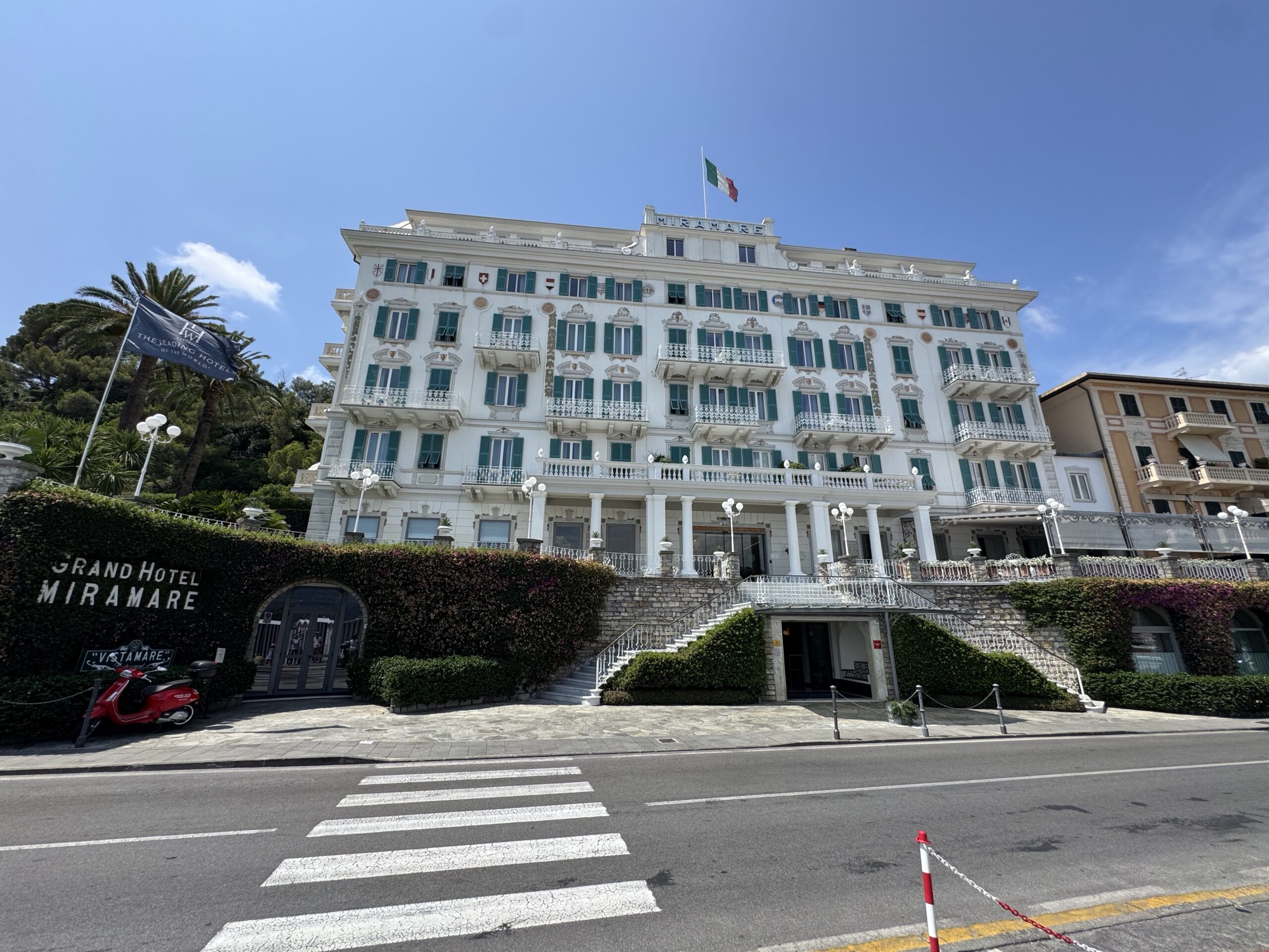 White grand hotel with green shutters, ornate balconies, and an Italian flag atop, facing a crosswalk and street below.