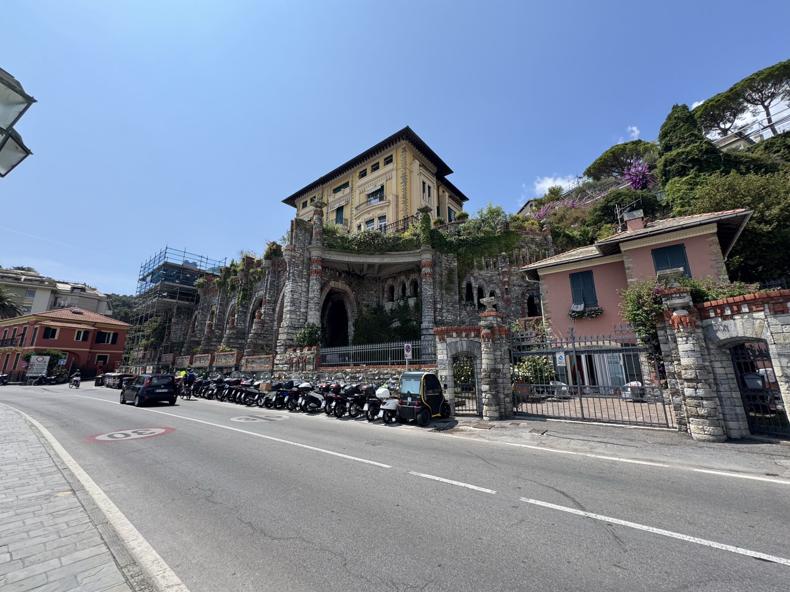Colorful hillside street with a historic stone building perched above arches, blue sky overhead and parked motorcycles along the curb.