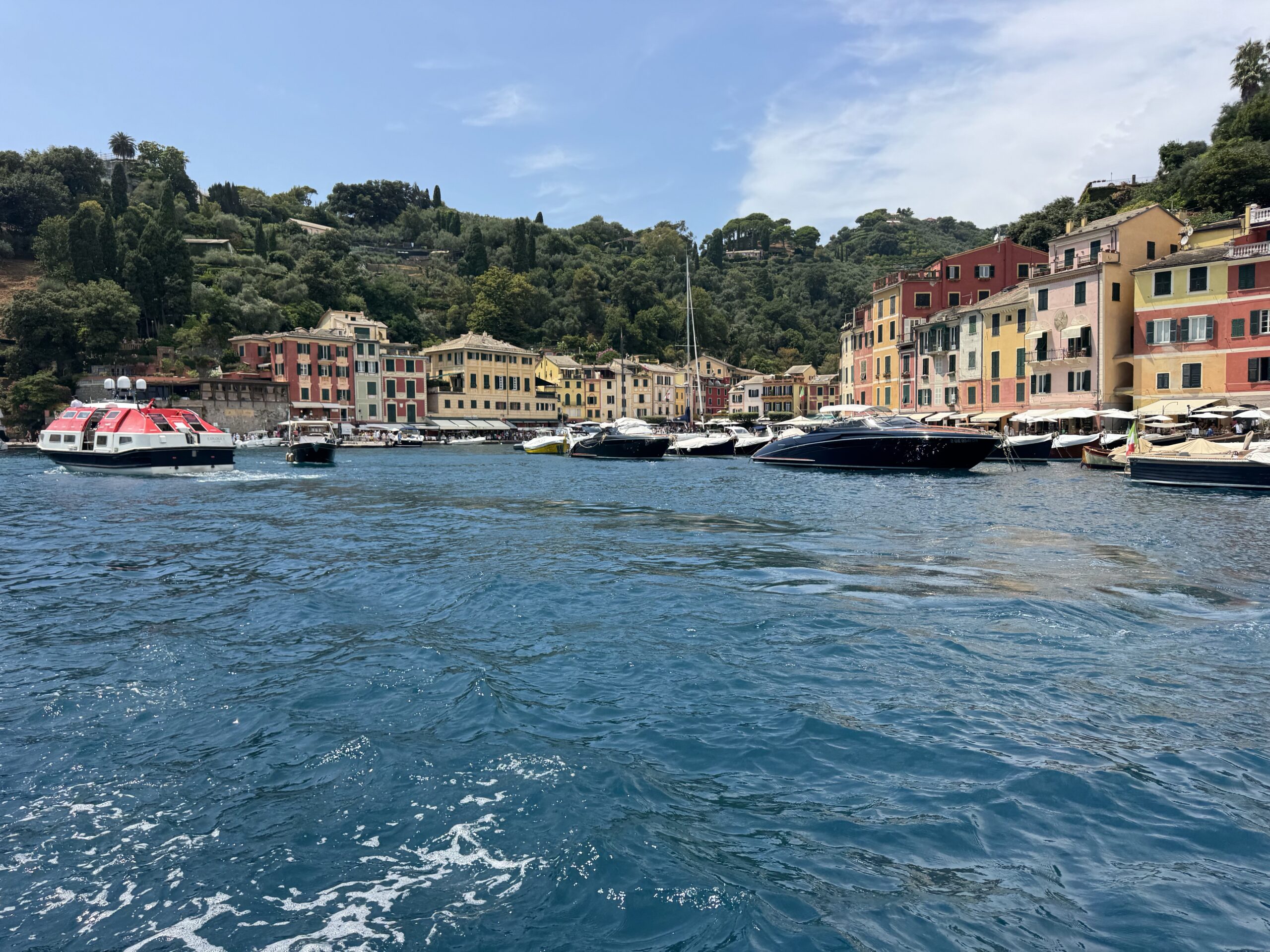 Colorful waterfront town with pastel buildings along a harbor and boats in the blue sea, hillside in the background.