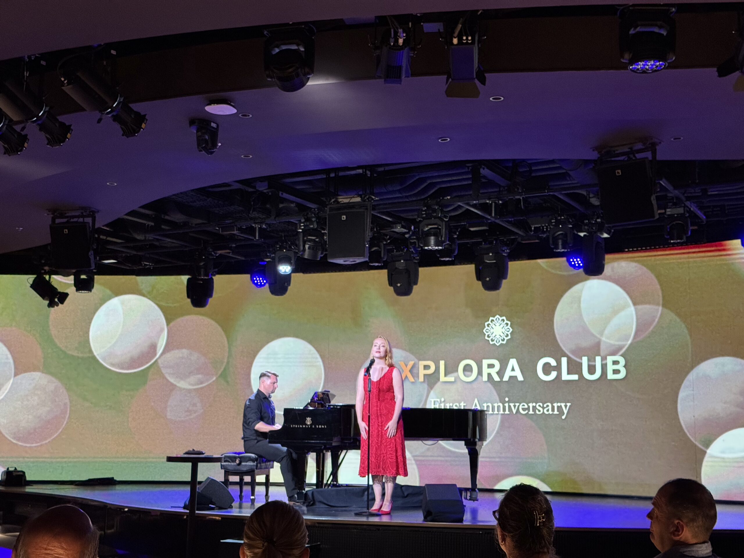 Stage performance: a pianist on a keyboard accompanies a female singer in a red dress, with an audience visible in the foreground and a colorful backdrop behind them.