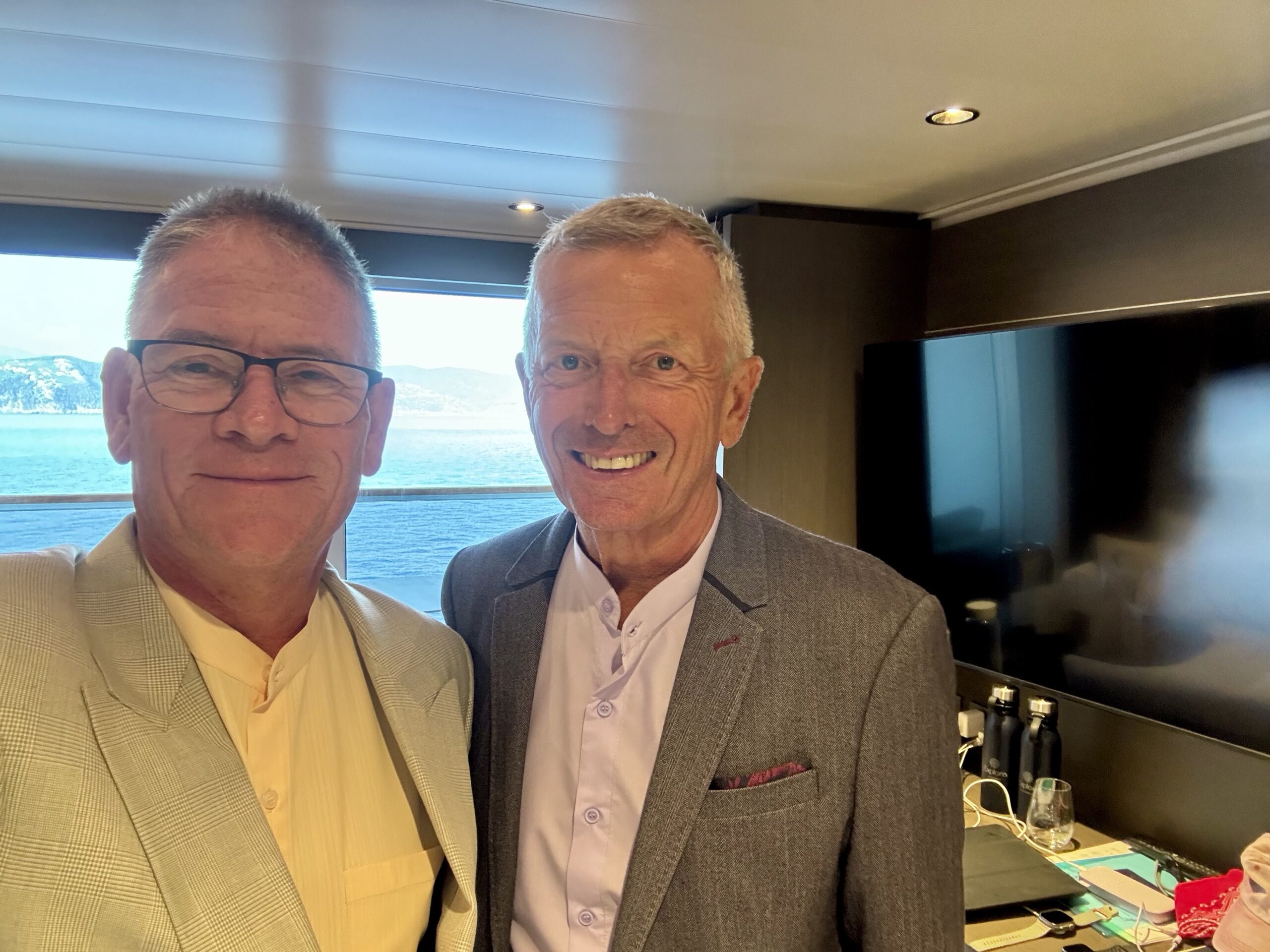 Two smiling men in suits posing for a selfie indoors with a sea view through the window behind them.