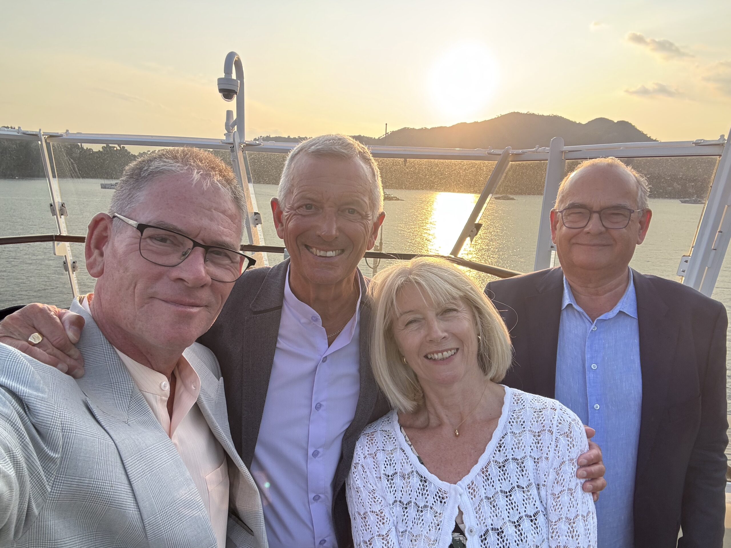 Group of four adults taking a sunset selfie on a boat, smiling with water and hills in the background.