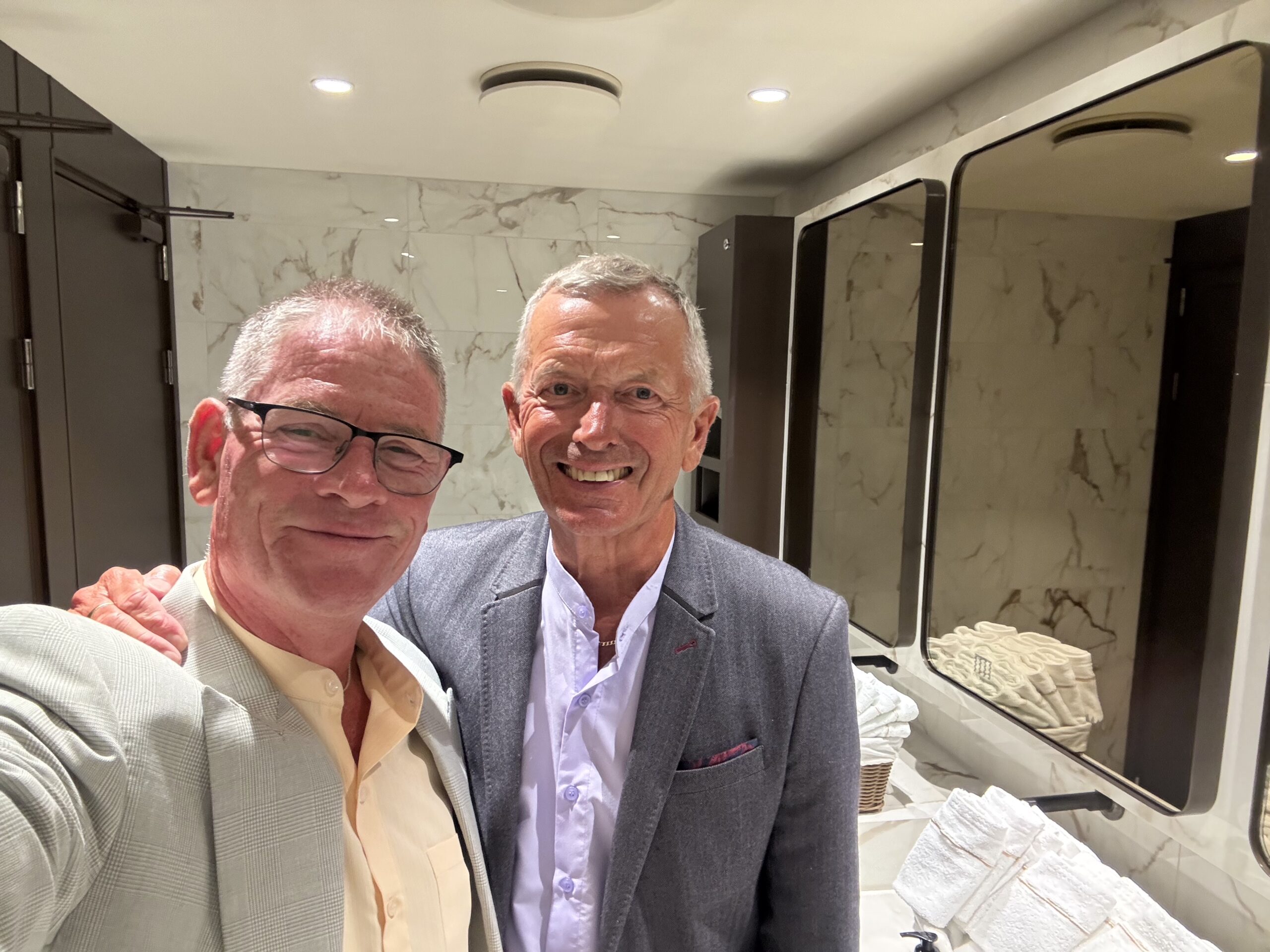 Two men in suits smiling while taking a selfie in a marble bathroom with large mirrors and towels nearby.