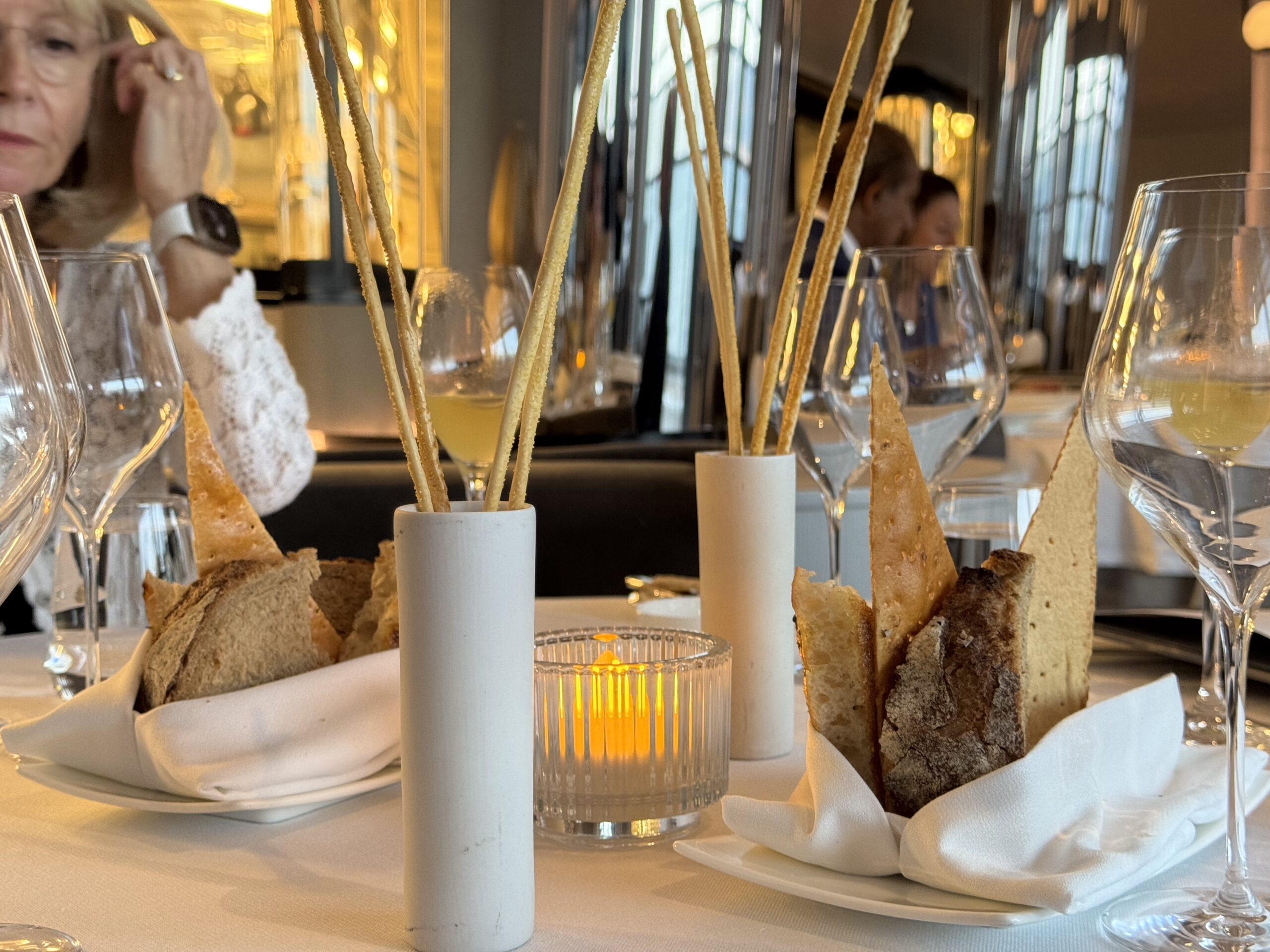 Restaurant table setting with breadsticks in white cylindrical holders, crusty bread, and lit candle in a glass holder; blurred diners in background.