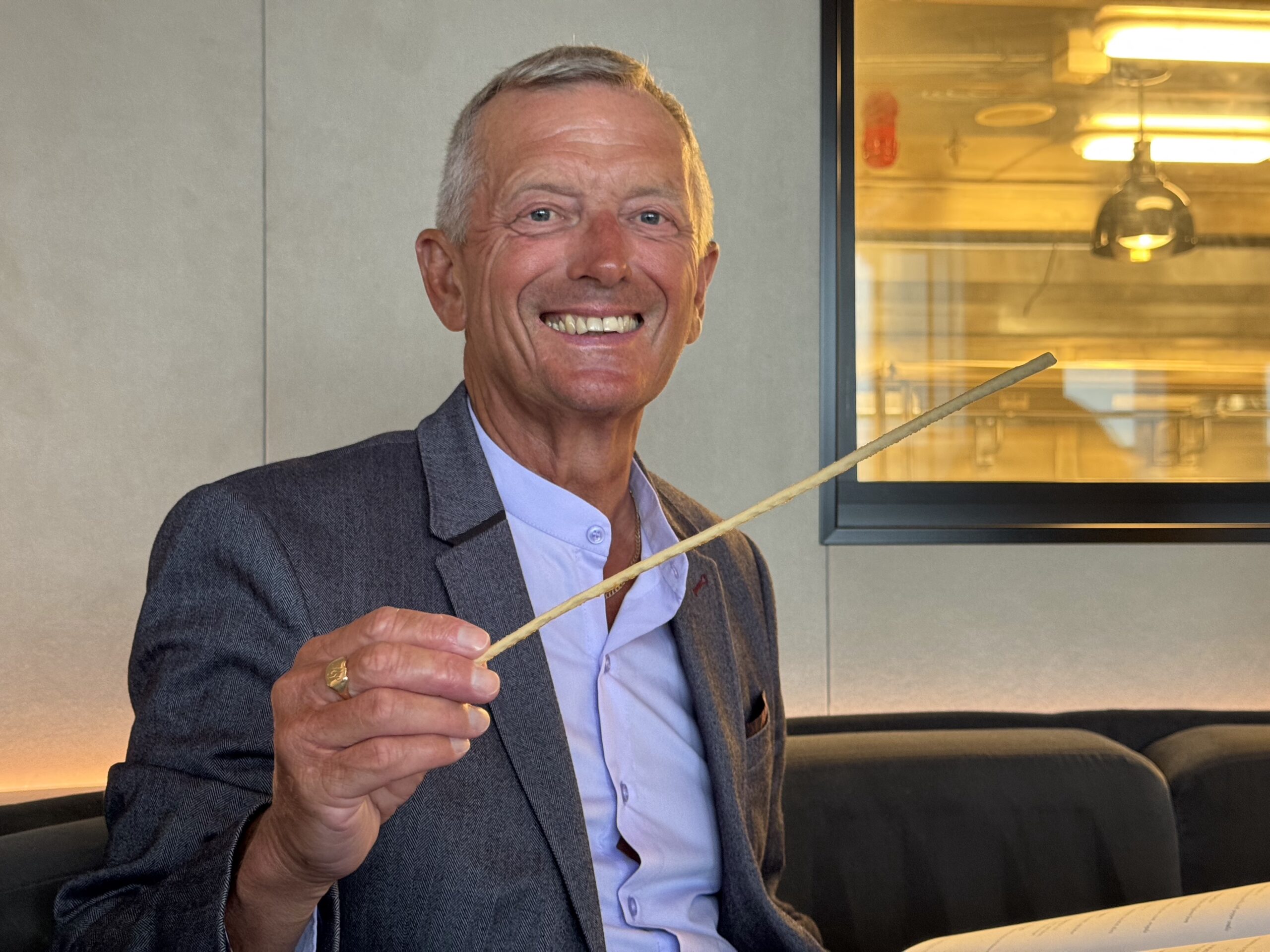 Smiling man in a gray blazer holding a wooden stick indoors, seated on a couch.
