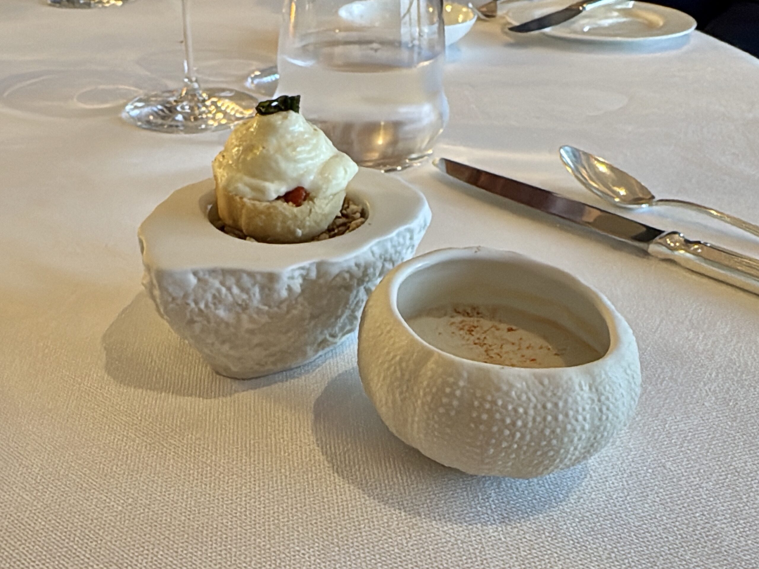 Small savory bite served in a white ceramic cup with a creamy topping and herb garnish on a restaurant table.
