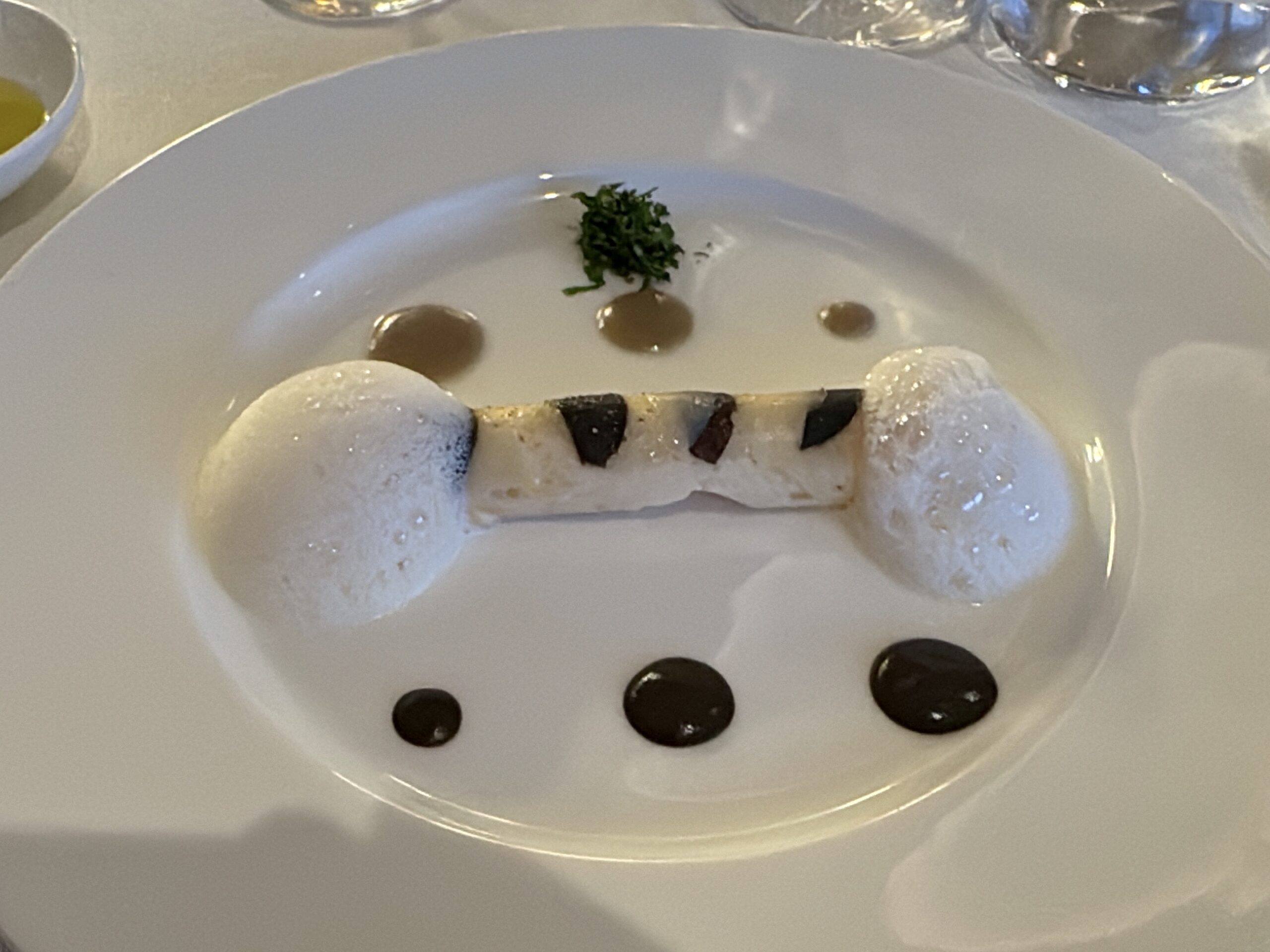 A white plate with a small green herb garnish in the center, several brown sauce droplets around, and white foam or meringue pieces at the edge.
