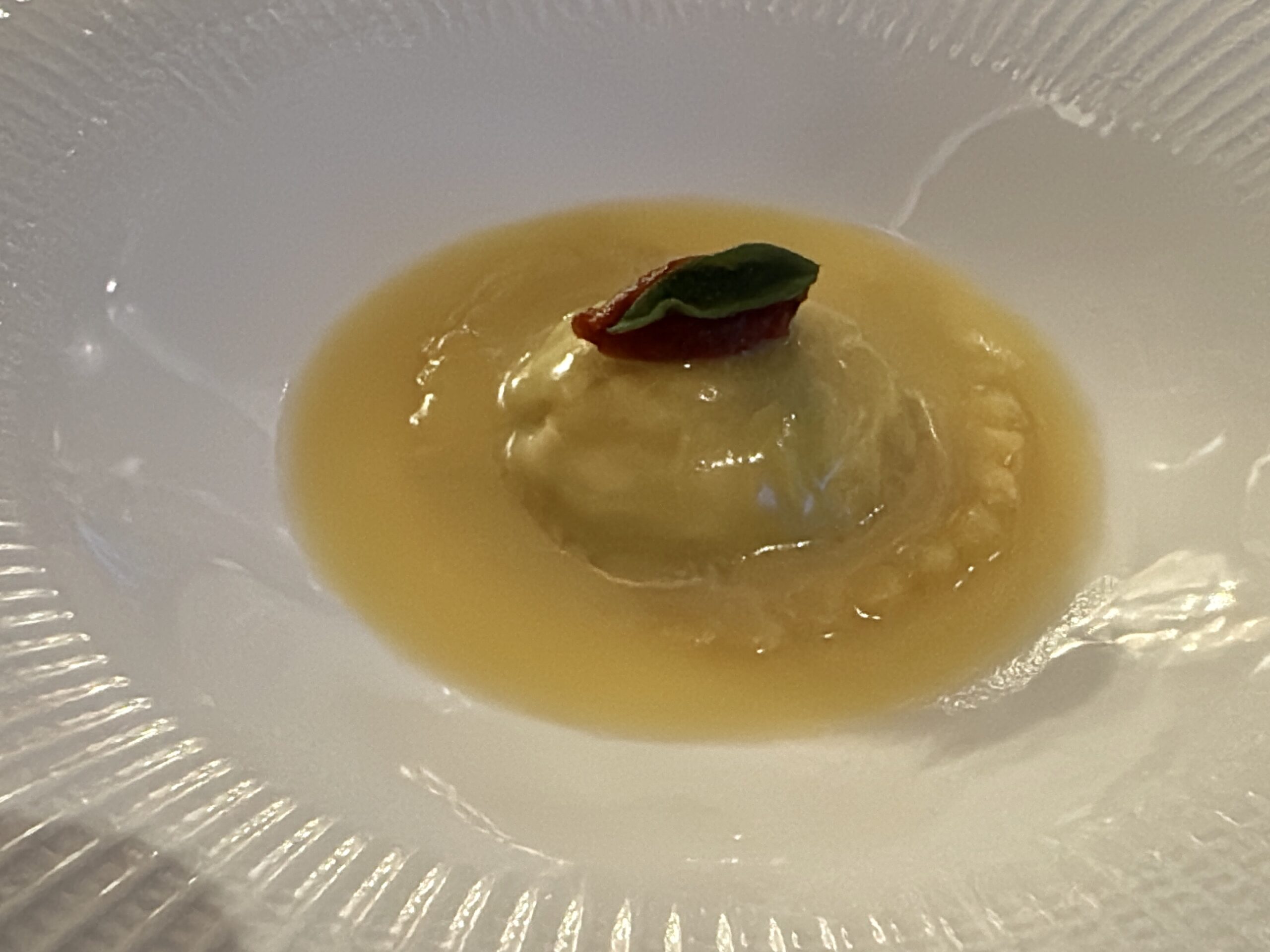 A plated dumpling in a pale yellow sauce, topped with a small green leaf and red garnish on a white plate