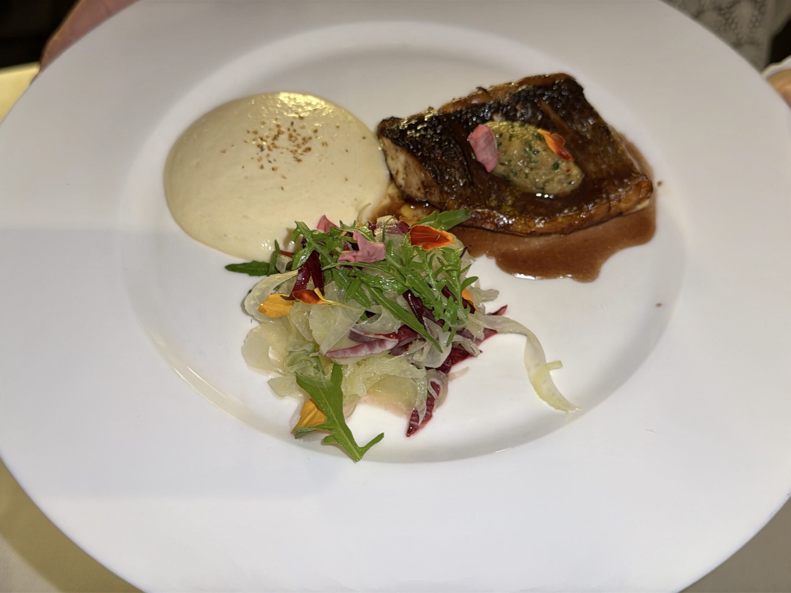 Seared meat in brown glaze with a side of pale creamy sauce and a colorful herb salad with edible petals on a white plate.