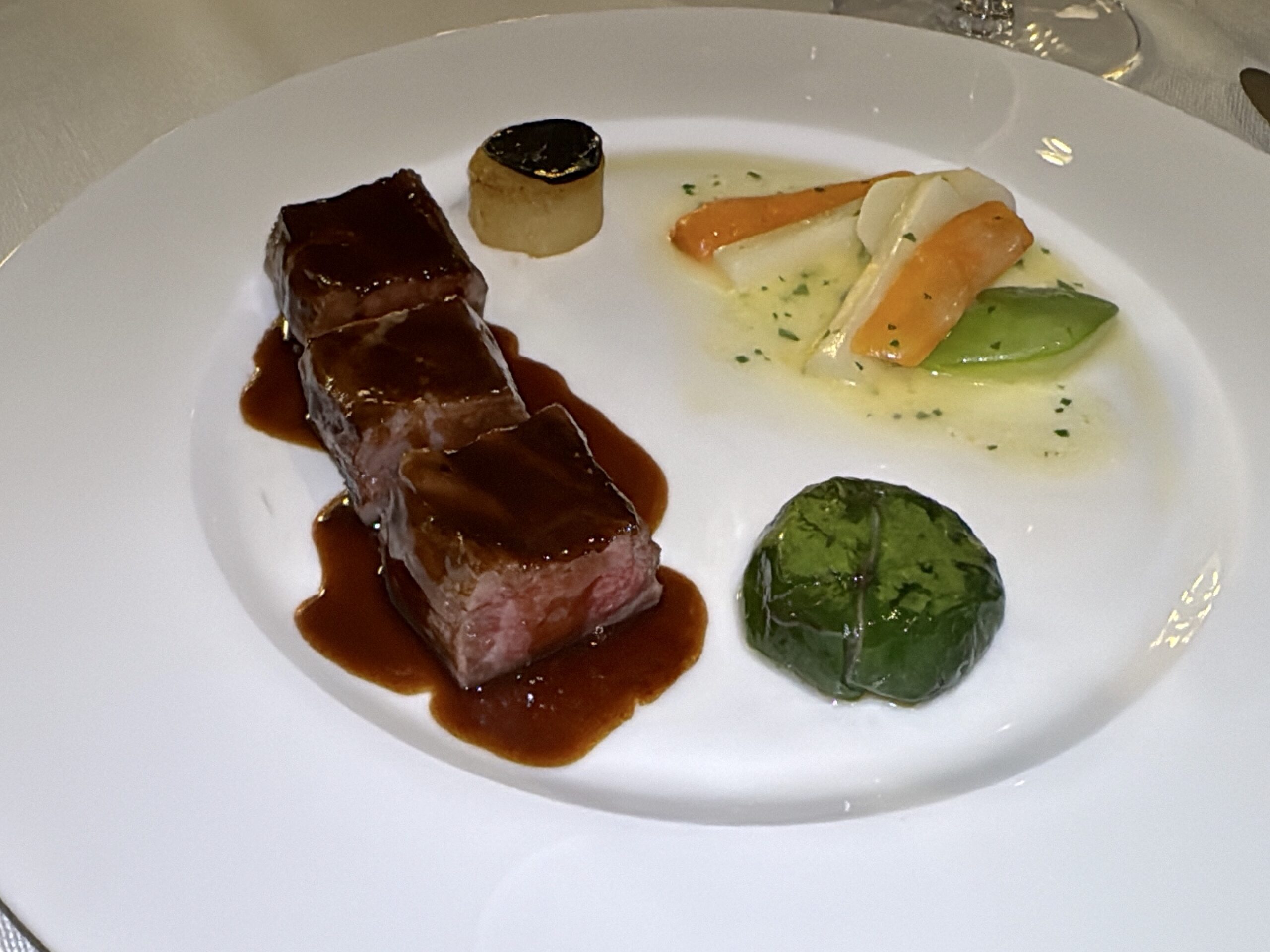 Beef tenderloin medallions in a rich brown sauce with roasted carrot and white asparagus beside a spinach-wrapped vegetable on a white plate.