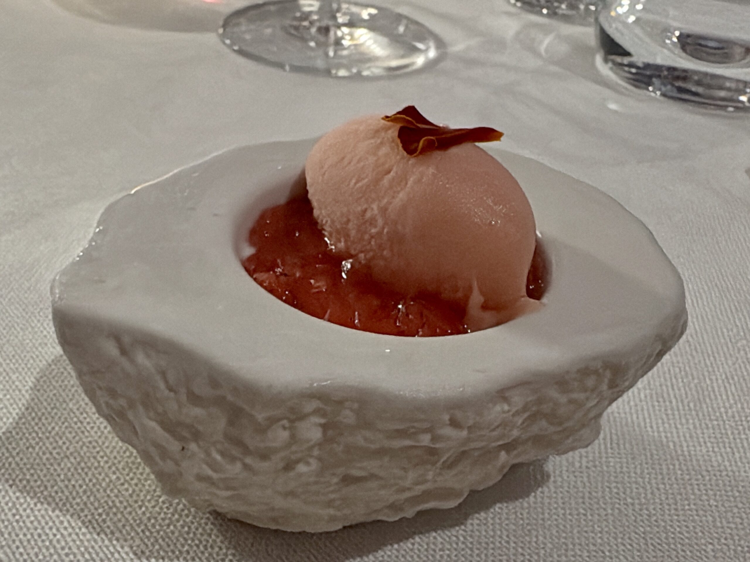 Close-up of a pink sorbet scoop topped with a small orange flower petal on a white plate with sauce nearby, dining setting visible in the background.