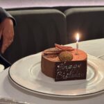 Chocolate birthday cake with a single lit candle and decorative citrus slices on a white plate covered by a silver charger.