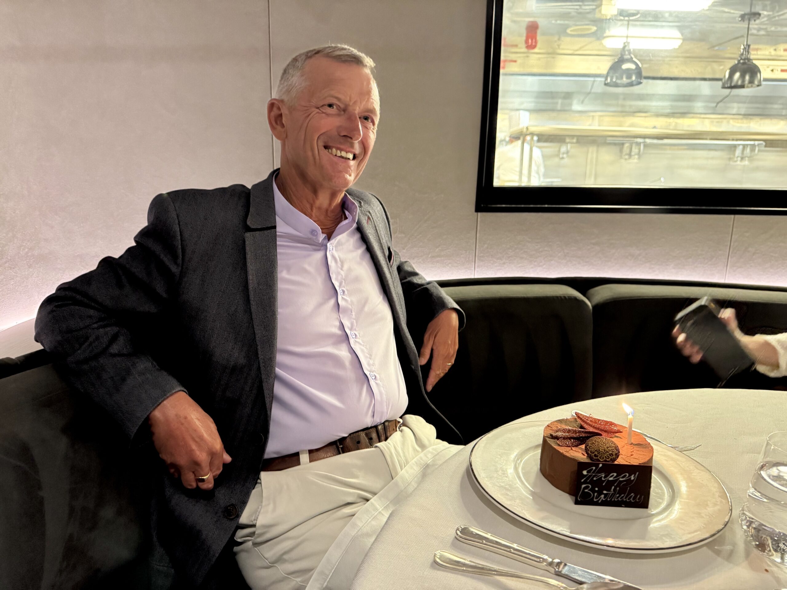 Smiling man in a blazer sits at a table with a chocolate birthday cake and candle in front of him.