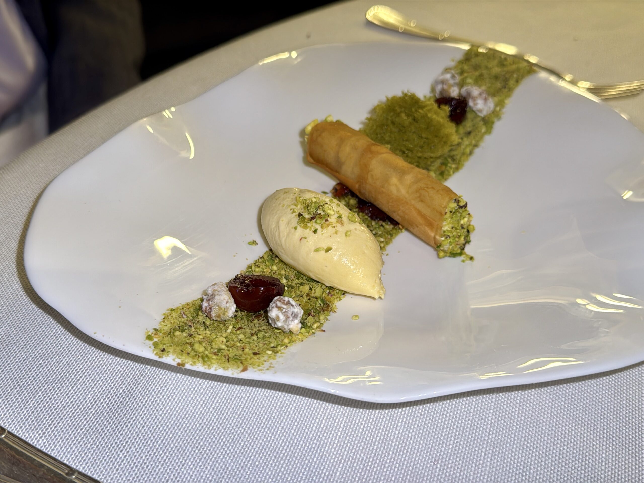 Dessert plate with a pistachio-crusted pastry roll, a scoop of vanilla ice cream, and green pistachio crumbs on a white plate.
