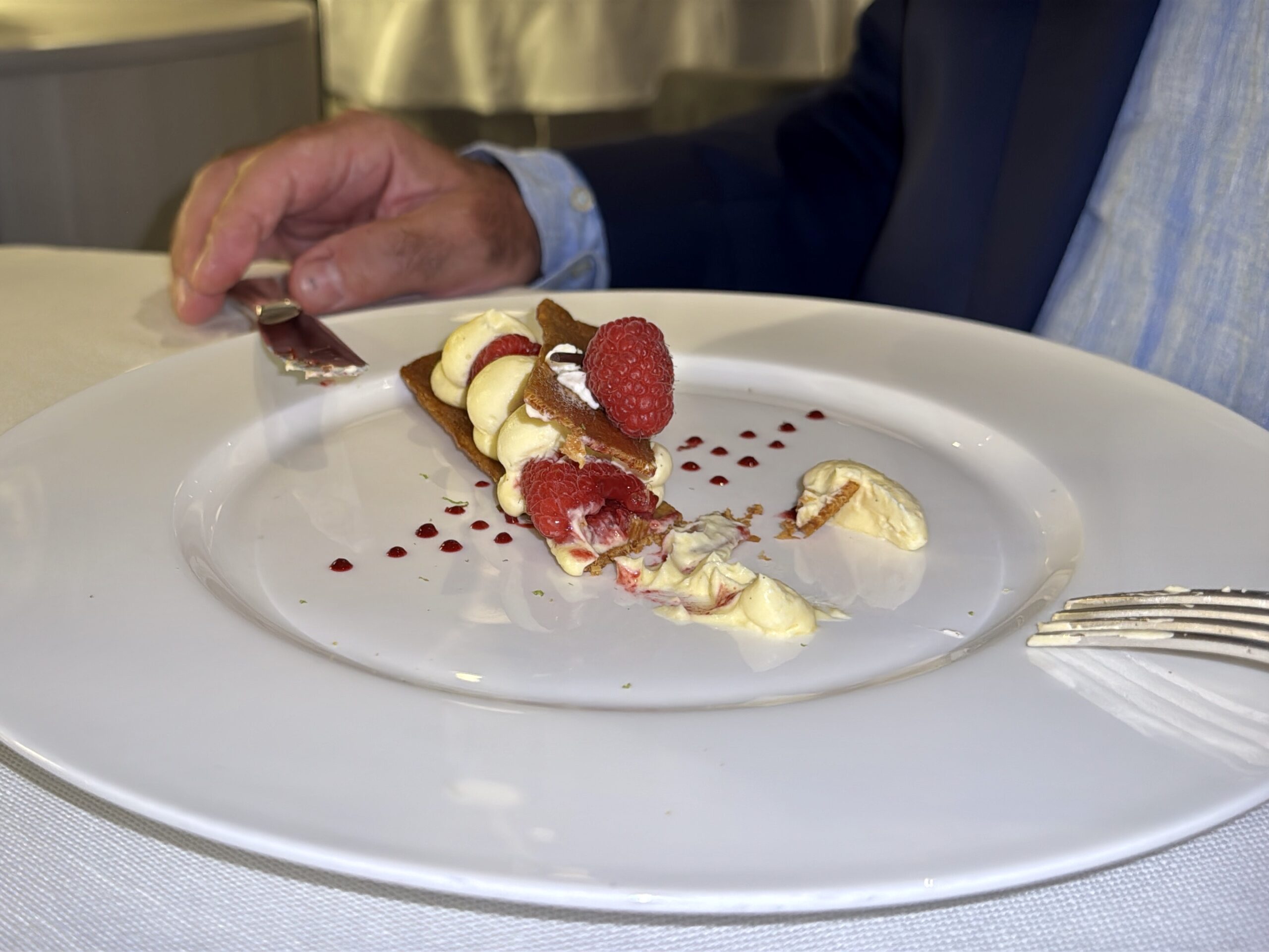 A hand in a suit cuts into a raspberry-topped pastry with cream on a white plate.