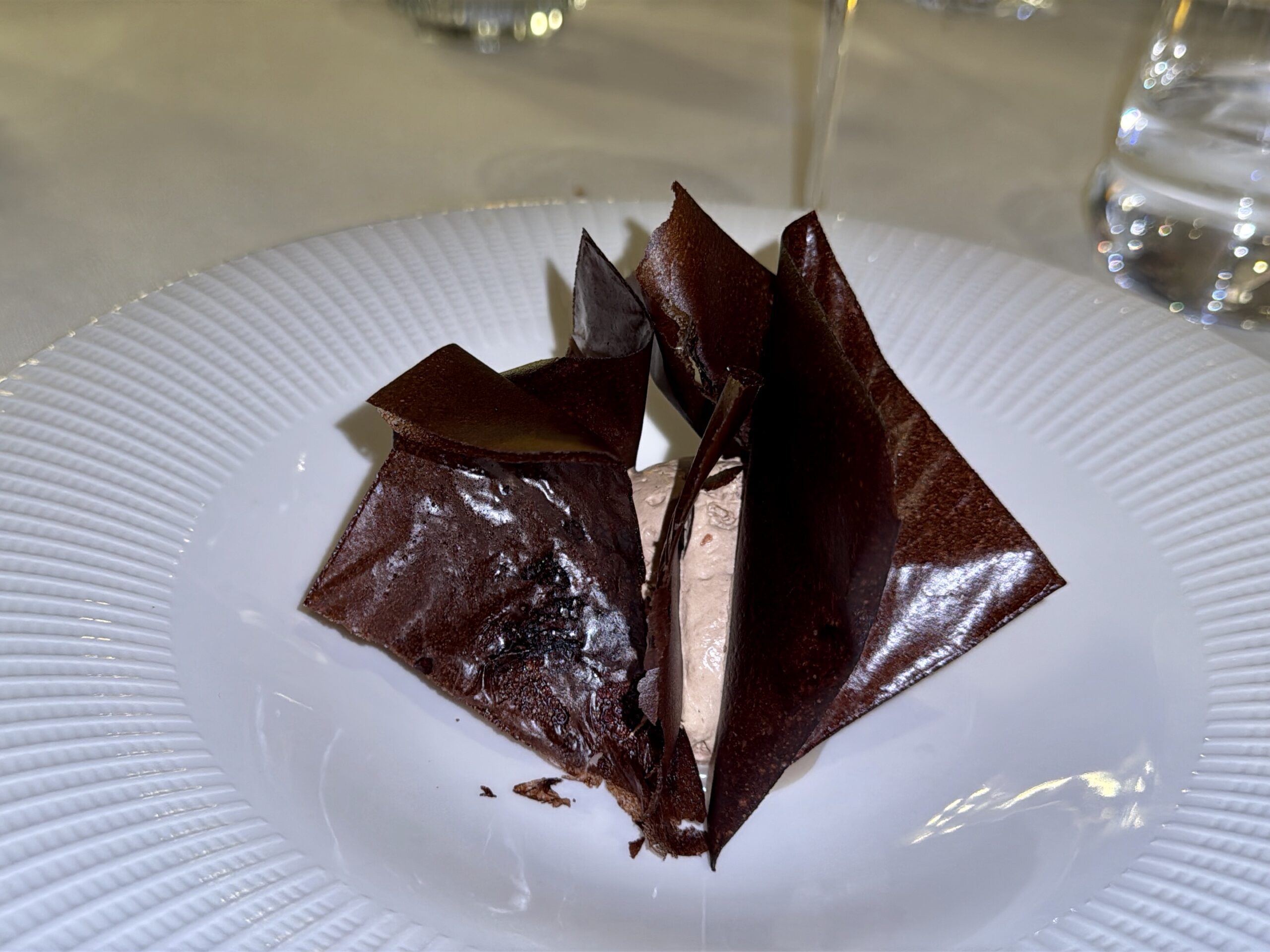 Dessert plate with dark chocolate shards arranged like a flower around a light brown mousse. A glass is blurred in the background.
