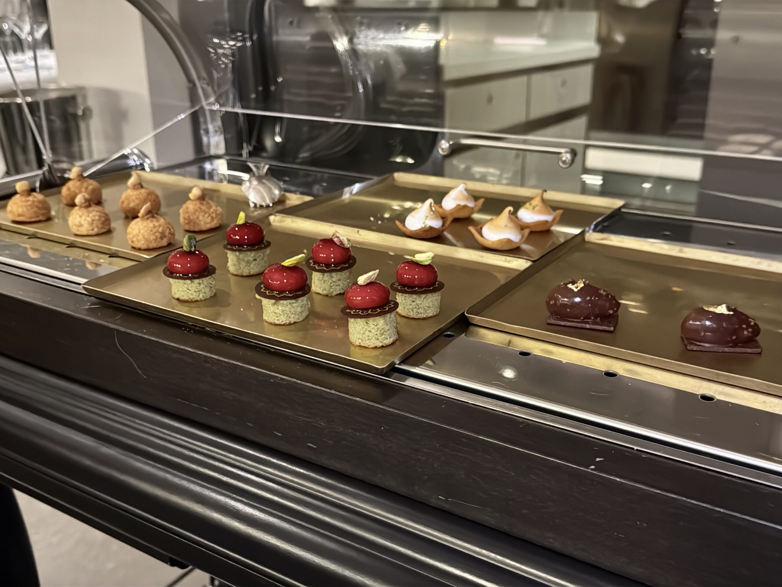 Assorted pastries on metal trays inside a glass bakery display case, including cream puffs and red glazed desserts.