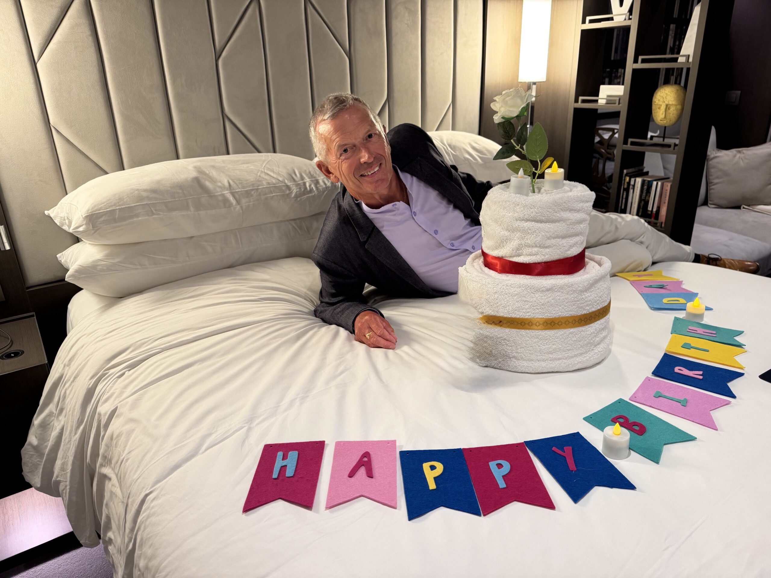 Man in a suit lying on a bed, smiling, with a towel cake topped with a rose and candles nearby.